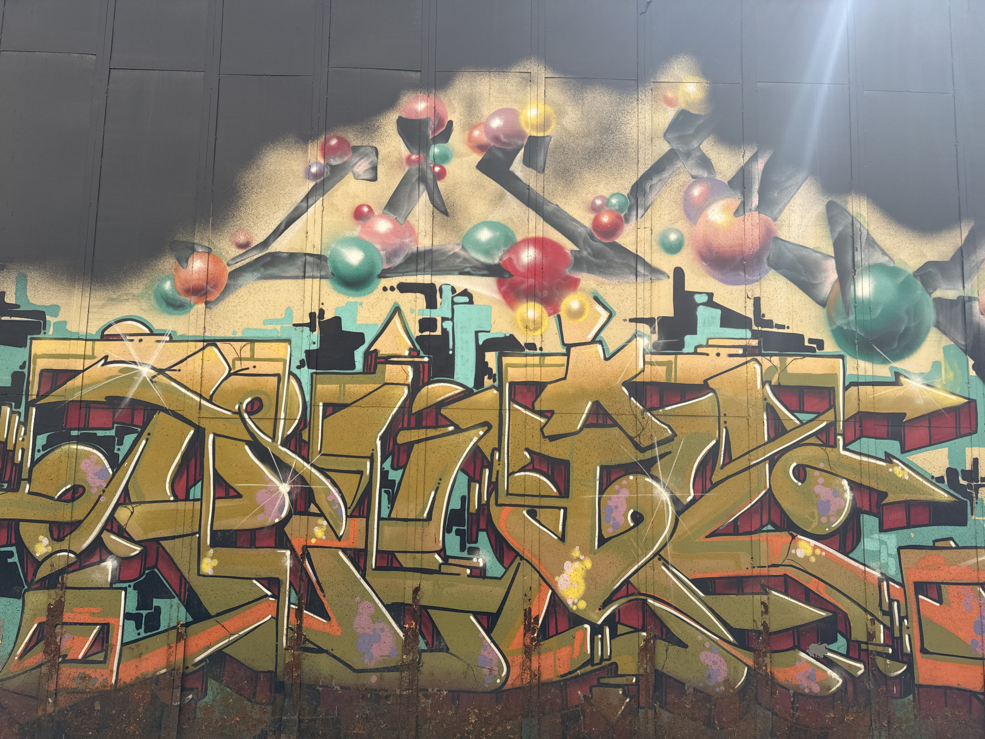 The image depicts a section of a wall covered in graffiti. The wall appears to be constructed of large, vertical panels. The upper portion of the wall is painted a dark gray color, transitioning into a tan or beige background.
The graffiti features a complex design with overlapping shapes and colors, including shades of brown, green, orange, blue, red, and black. Abstract shapes and lines are used to create a stylized effect. Above the main text are black shapes with colored spheres.
The bottom portion of the wall is weathered and shows signs of rust or corrosion. The style of the graffiti suggests urban art.
The photo was taken in Moscow, Russia.