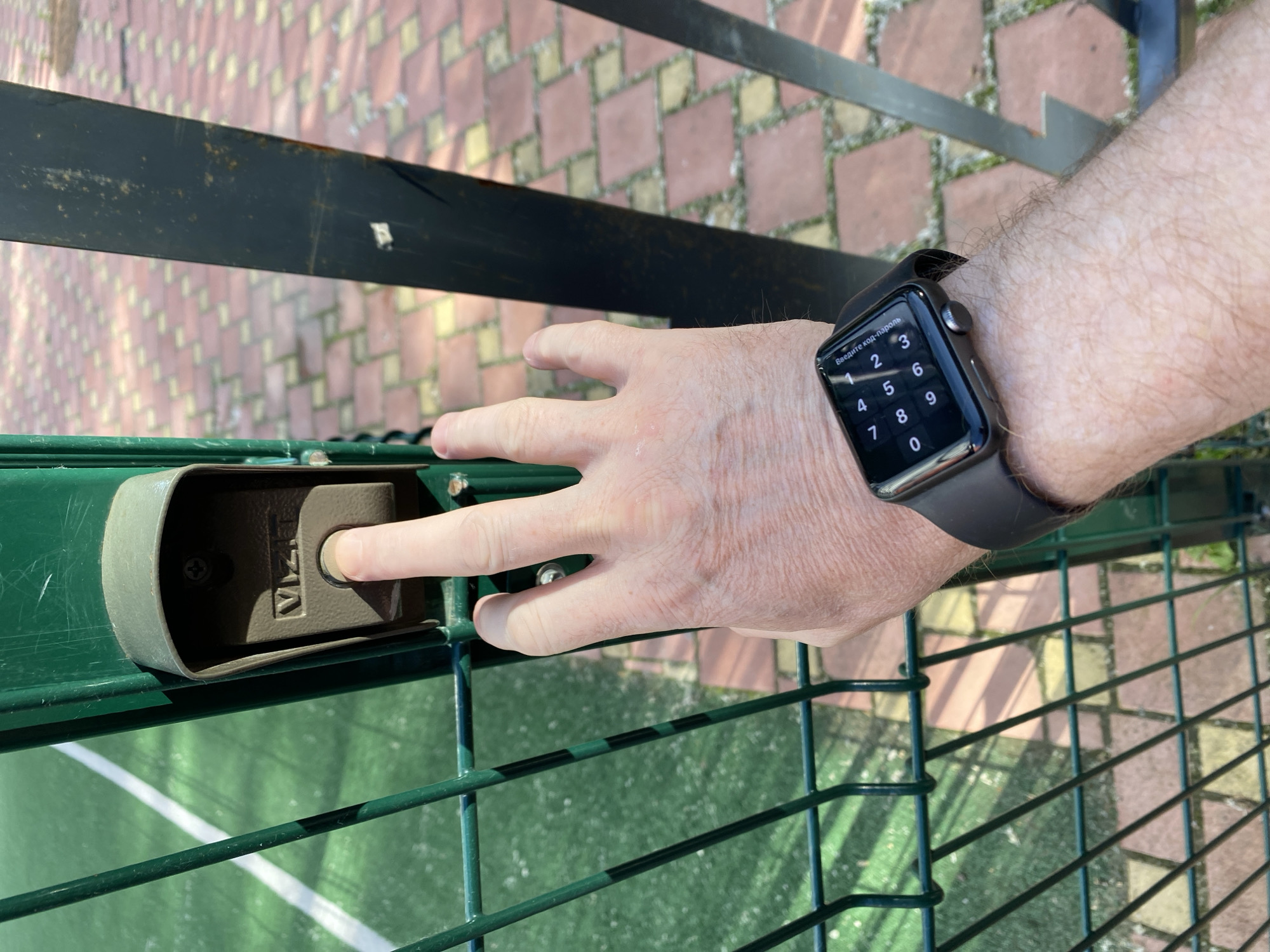 A close-up, eye-level shot shows a person's right hand pressing a button on an intercom system mounted on a green metal fence post. The intercom is a gray, oval-shaped device with the word "VIZIT" clearly visible. The hand is bare, and the person is wearing a dark-colored smartwatch on their left wrist. The watch face displays a numeric keypad for entering a code or password. The background features a green, outdoor court surface with white lines, possibly a tennis or basketball court, and a patterned brick pathway. The image appears to have been taken outdoors in daylight.