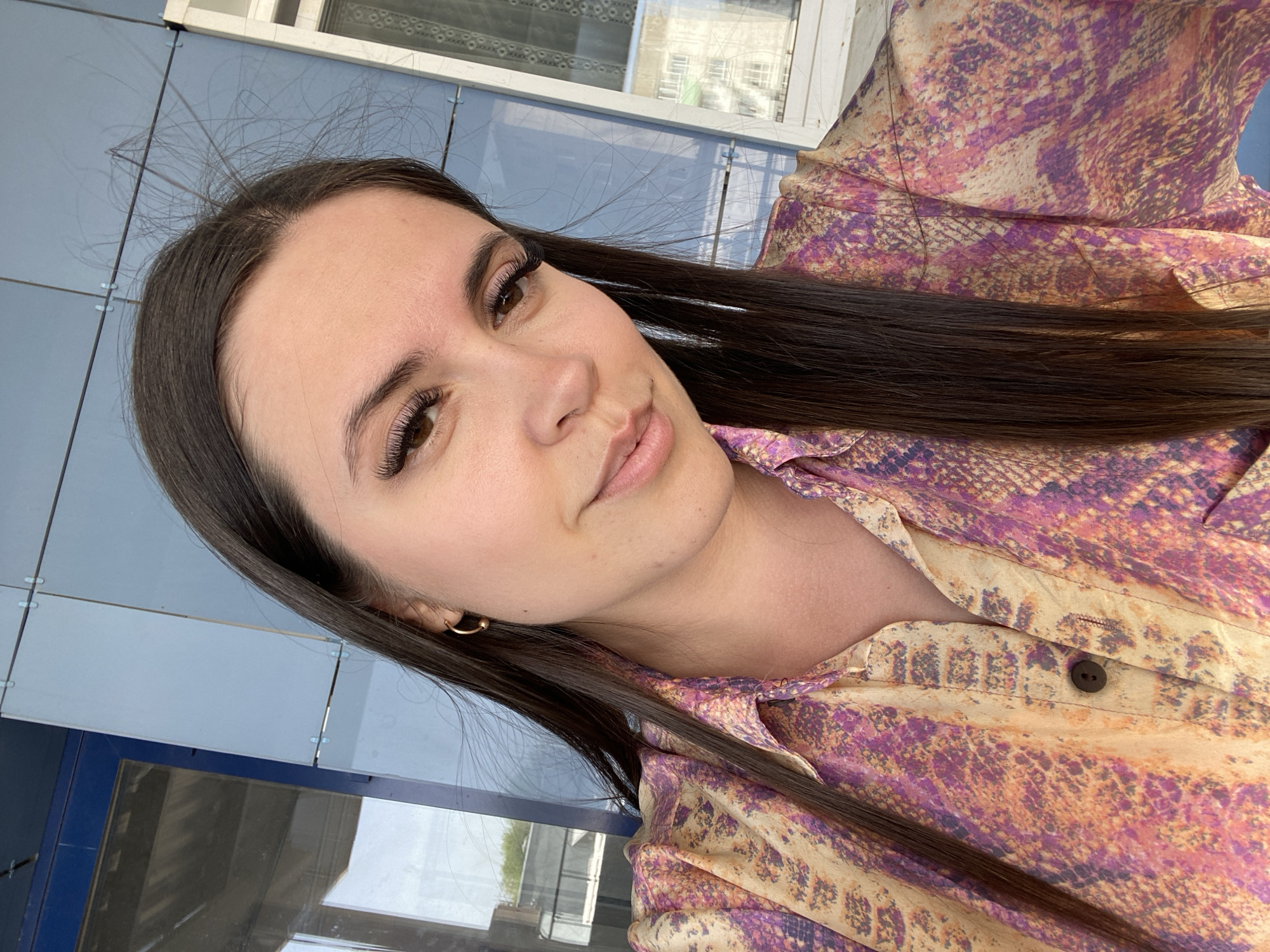A young woman with long brown hair and prominent false eyelashes poses for a selfie outdoors. She wears a collared shirt with a snakeskin-like pattern in shades of purple, pink, and beige. A small gold hoop earring is visible in her left ear.
The background consists of a blue-grey panelled exterior wall with a window above, and a lower section with reflective surfaces. Based on the urban architectural style visible in the background, the location is likely within the city of Chaikovsky, Russia.