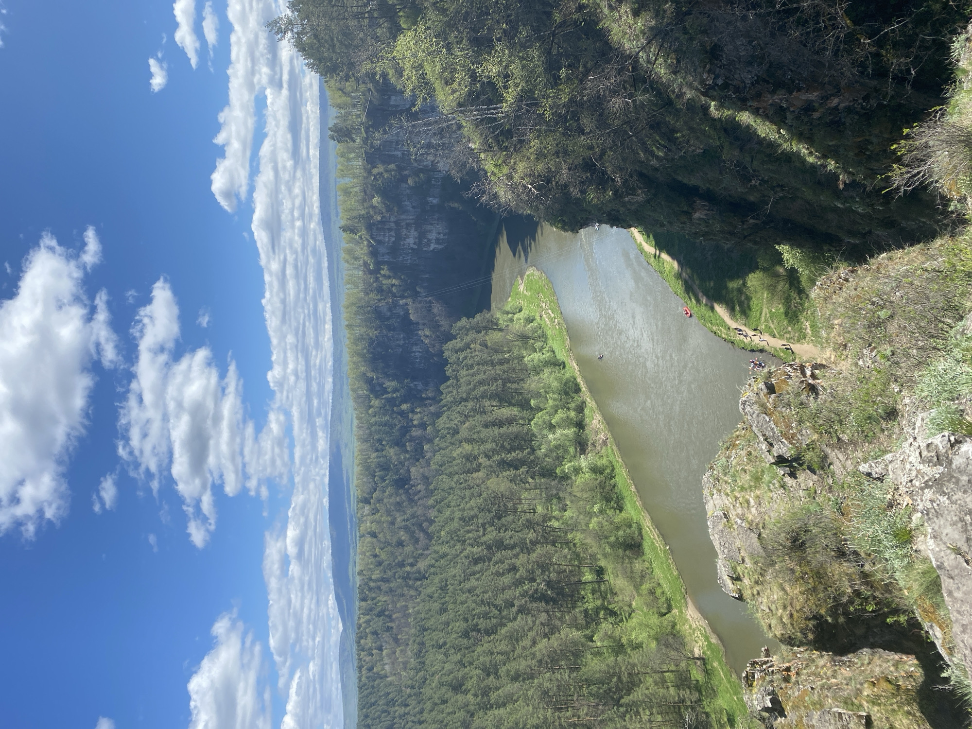 The image shows a river cutting through a forested area in Alekseevka, Russia. The perspective is from a high vantage point, possibly a cliff edge, with rocky terrain in the foreground. The river appears relatively calm, with hints of a red boat on the water. A narrow strip of land with a pathway borders the river. A group of people are standing near the edge of the cliff where the river bends. The sky is blue with scattered white clouds, and a forest stretches into the distance along the horizon.