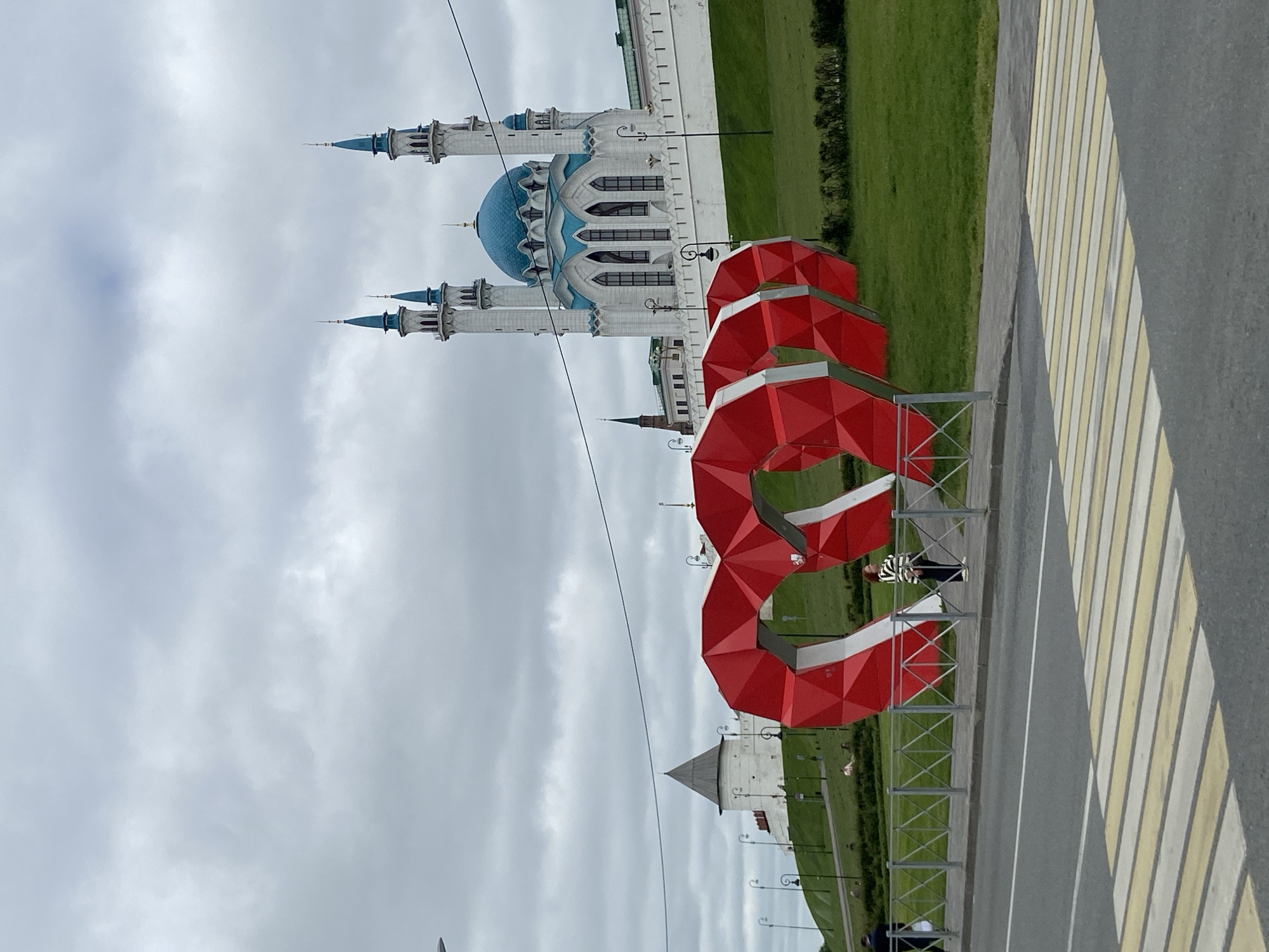 This image captures an outdoor scene featuring the prominent Kul Sharif Mosque in Kazan, Russia. The mosque, situated on the right side of the frame, is a grand, white structure with a large, sky-blue central dome and four slender minarets, each capped with a smaller blue spire. Its facade showcases intricate architectural details, including multiple arched windows with blue accents. Lush green grass surrounds the mosque, extending into the background.
In the foreground, a series of large, red, geometric sculptural elements with white accents are arranged along a landscaped grassy area. These abstract structures stand behind a low metal fence, which separates the green space from a paved road. The road features a distinct yellow and white striped crosswalk. One person, wearing a striped shirt and dark trousers, is visible behind the red sculptures, near the fence. The sky above is overcast with a uniform layer of grey clouds, indicating a cloudy daytime. No discernible text is visible within the image.