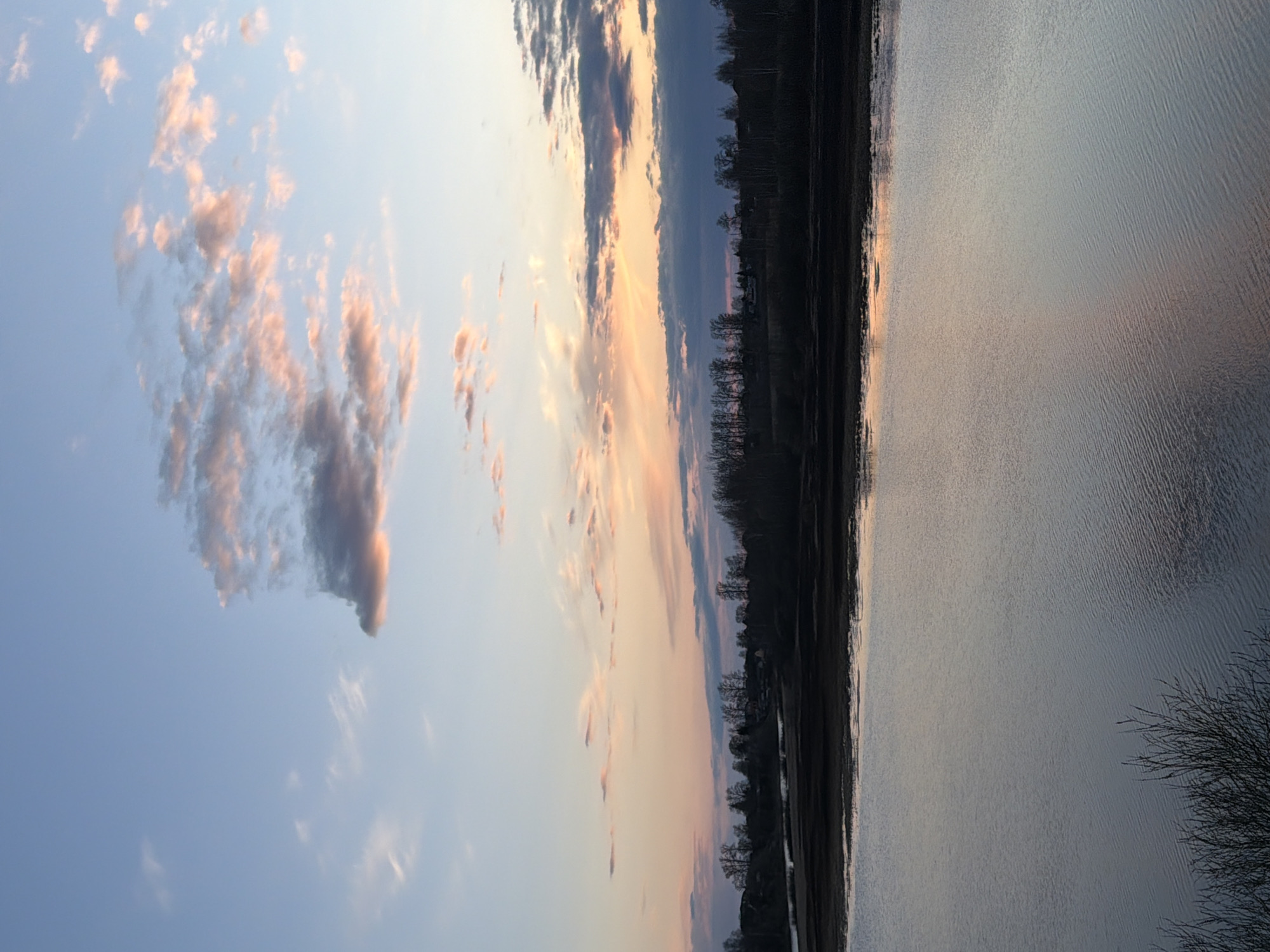 A serene horizontal view shows a body of water, likely a lake or river, reflecting the colors of the sky. The sky transitions from a clear blue to hues of orange and pink, suggesting a sunset or sunrise. Clouds are scattered across the sky, some with pinkish tinges. A dark treeline runs along the edge of the water, separating it from what appears to be land. The location cannot be determined as the city is unknown.