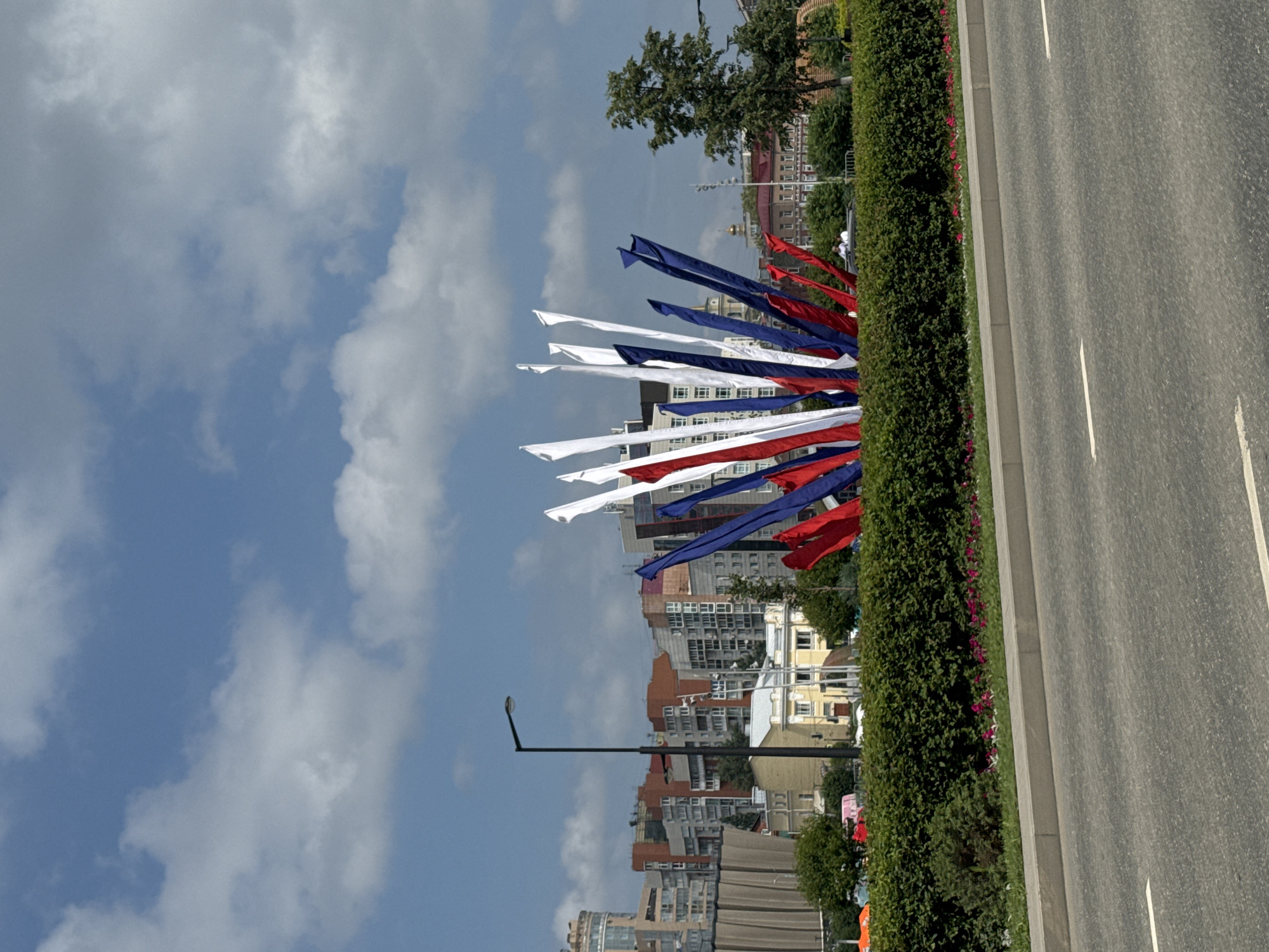 In the city of Perm, Russia, on a bright, partly cloudy day, a vibrant display of red, white, and blue fabric flags or streamers juts upwards from a meticulously trimmed green hedge. These colorful elements are arranged in a fan-like shape, creating a striking focal point. Behind the hedge, a paved road is visible, bordered by a concrete curb. Further back, the urban landscape unfolds with a variety of buildings, ranging from multi-story apartment blocks with modern architecture to older, more traditional structures. A single black lamppost stands tall amidst the cityscape. A lone tree with dark green foliage is situated on the right side of the frame, complementing the urban backdrop. In the distance, a golden dome is faintly visible, suggesting the presence of a church or other significant religious building.