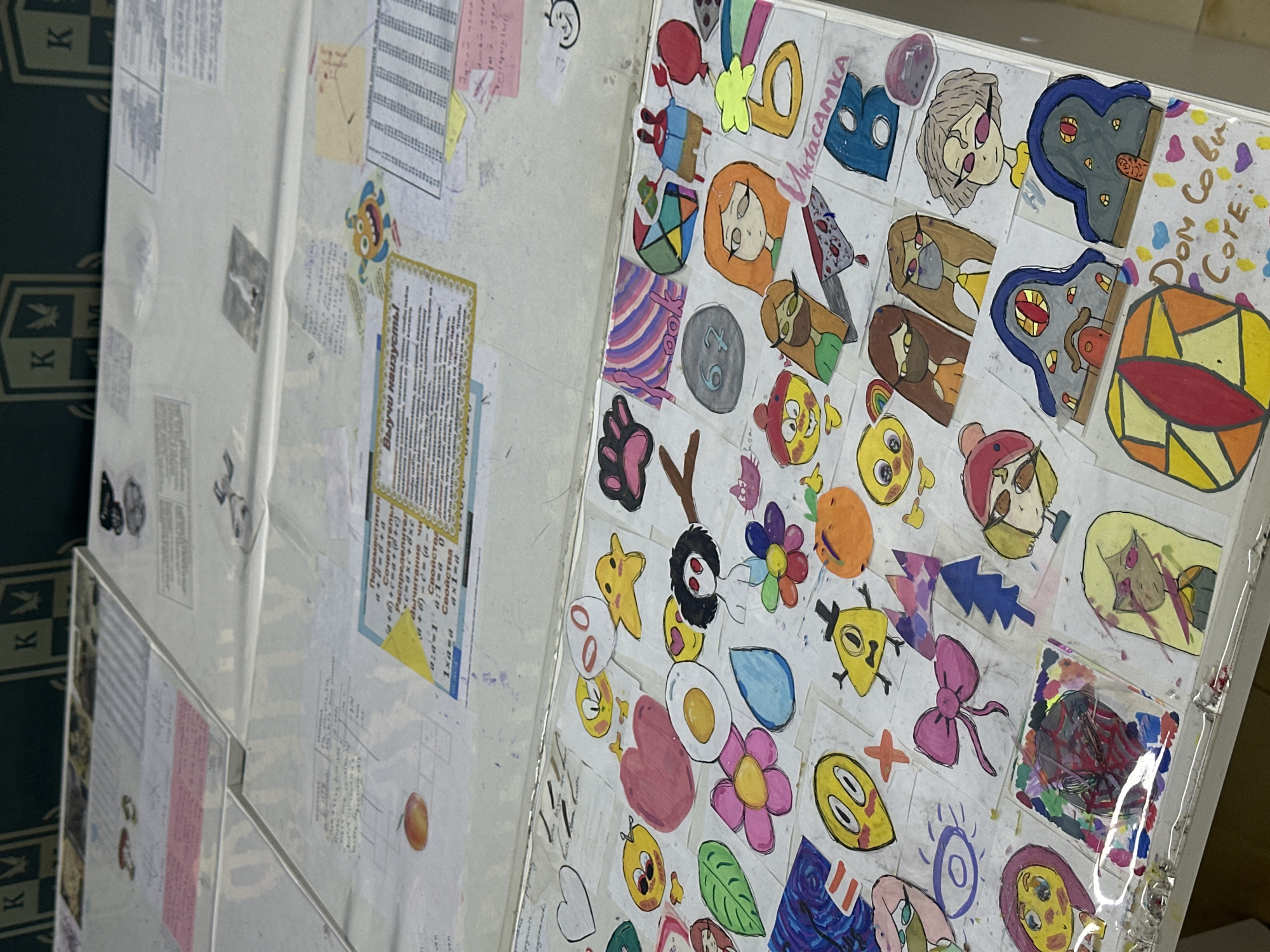 This image, taken in Sochi, Russia, showcases two adjacent decorated surfaces, likely part of a study area or personal display. The surface on the left is covered with various documents and papers, some pinned or taped down. These documents contain Cyrillic text, including what appears to be mathematical properties ("Свойства", "ax1-", "1xa=a") and a prominent yellow-bordered instruction "Выучи наизусть!" (Learn by heart!). Other less legible texts are also in Cyrillic, suggesting an educational context.
The surface on the right is a vibrant collage of numerous hand-drawn and colored cutouts or stickers. These colorful designs feature a variety of cartoon-style characters, emojis, and objects such as a paw print, a fried egg, flowers, hearts, a rainbow, and a bow. Notable characters include what appears to be Bill Cipher from Gravity Falls and figures reminiscent of Matryoshka dolls. Some stickers also contain text or numbers like "67," "B," "ИнdaCAMKA" (possibly a stylized "Instasamka"), and "Дом Сови Core" (likely referencing "The Owl House"). The stickers are arranged densely, creating a lively and personal decorative space.
The background on the far left reveals a dark fabric with a repeating 'K' logo, possibly from a uniform, bag, or furniture. The overall scene captures a personal and creative environment, combining elements of study with artistic expression. The lighting is even, suggesting an indoor setting.