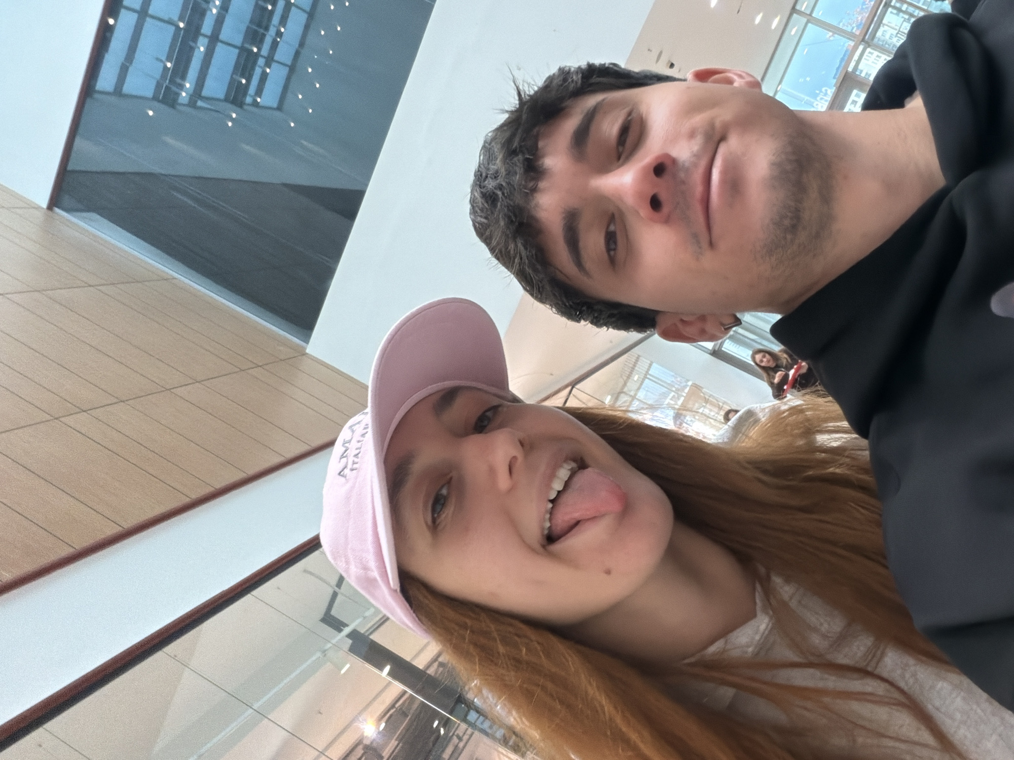 An upward-tilted selfie captures a young man and woman in an indoor public space, likely a modern shopping mall in Sofia, Bulgaria.
In the foreground, the woman dominates the lower half of the frame. She has long, reddish-brown hair cascading down her shoulders and wears a light pink baseball cap with the text "AMIA ITALIA" subtly visible on its front. Her face is turned towards the camera with a playful expression, her tongue sticking out and a wide, open-mouthed smile showing her teeth. She is wearing a light-colored top.
Behind and slightly to the right, the man is partially visible. He has dark, short hair and stubble on his chin and jawline. He is dressed in a black hooded top and looks towards the camera with a neutral, slightly smirking expression.
The background reveals a brightly lit, contemporary interior. Large glass windows reflect numerous small, bright lights from the ceiling or opposite walls, creating a shimmering effect. Parts of light-colored walls and light wood-paneled flooring are visible, consistent with a modern commercial or public building. The overall lighting suggests it is daytime. The image conveys a casual and lighthearted moment between the two individuals.
