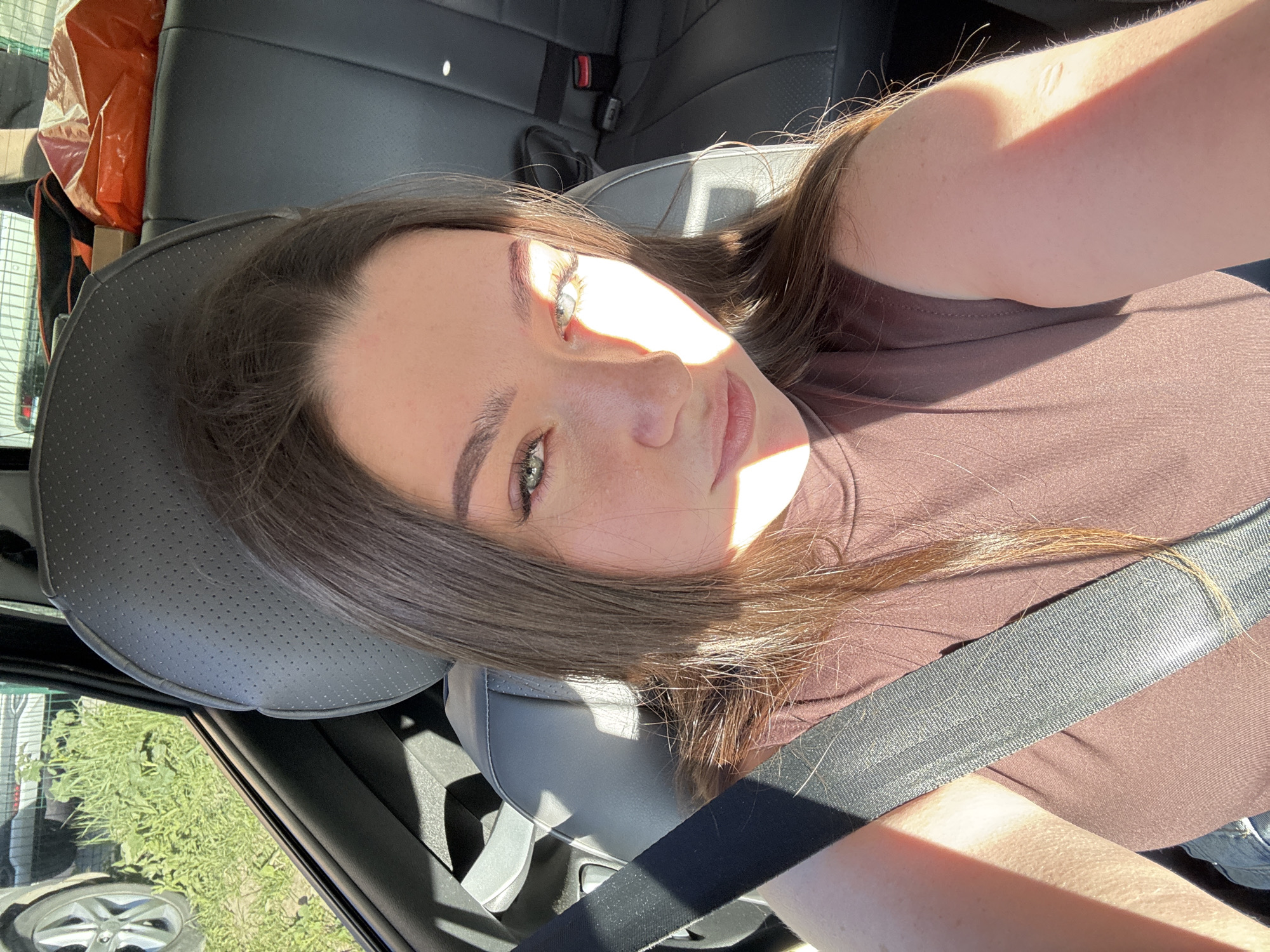 A woman with brown hair and green eyes is in the driver's seat of a car. She is wearing a brown shirt and a seatbelt. The car is parked and there are buildings visible through the window. The location of the image is not explicitly stated as Perm, Russia, nor is any other precise location, therefore I cannot identify the precise location.