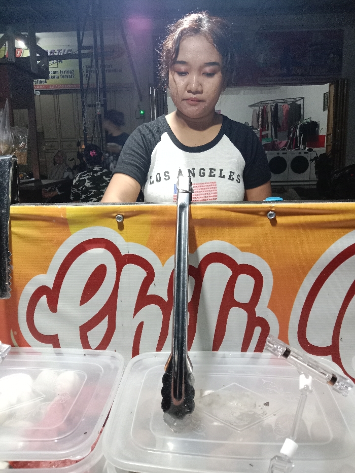 A woman of apparent Southeast Asian descent stands behind a commercial counter in Turunsih, Indonesia. She wears a white raglan-style t-shirt with black sleeves, featuring "LOS ANGELES" and an American flag-like graphic. Her hair is dark and medium-length, and she gazes downwards and slightly to her left. The counter, fronted by an orange and white banner with red script, displays clear plastic containers holding white and pink spherical food items. A pair of silver tongs stands upright in one container, and a clear plastic syringe-like object lies on the counter to the right. In the background, artificial lighting illuminates adjacent open-front businesses. To the left, a general store or repair shop is visible with various signs. To the right, a laundry service featuring multiple washing machines and hanging garments is present, with a dark motorcycle parked nearby. Several indistinct individuals are visible in the background, some seated.