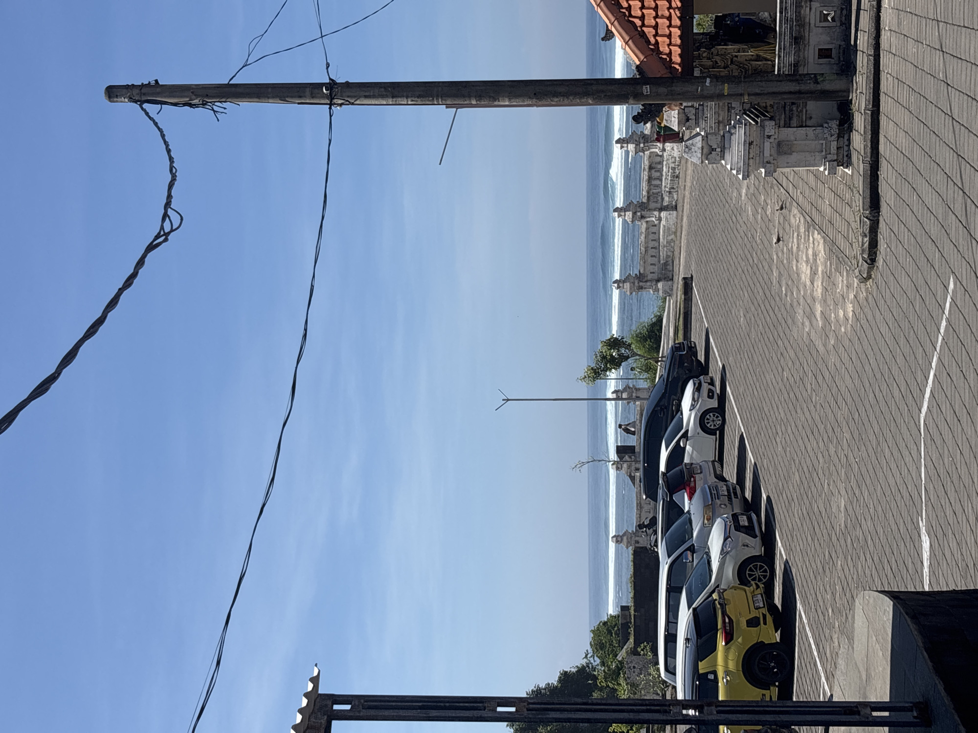 The image shows a view in Kuta Selatan, Indonesia. The scene is a parking area with cars lined up next to a traditional Balinese stone wall or structure. Beyond the wall, the ocean is visible with waves breaking. An electrical pole with wires runs along the left side of the frame. The sky is clear and blue. The picture appears to be taken from an elevated position.