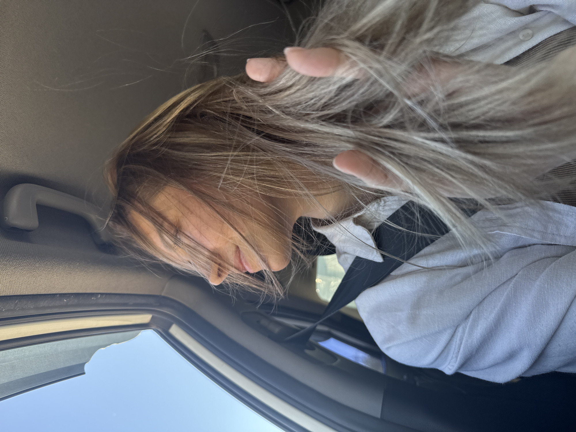 The photograph is taken from within a car, looking toward the passenger side. A fair-skinned person is leaning forward, their head tilted down and their blonde hair is partially covering their face and spread across the interior of the car. The person is wearing a white shirt and a seatbelt. A clear blue sky is visible through the car window. The location cannot be determined as Chaikovsky, Russia from the details in the image.