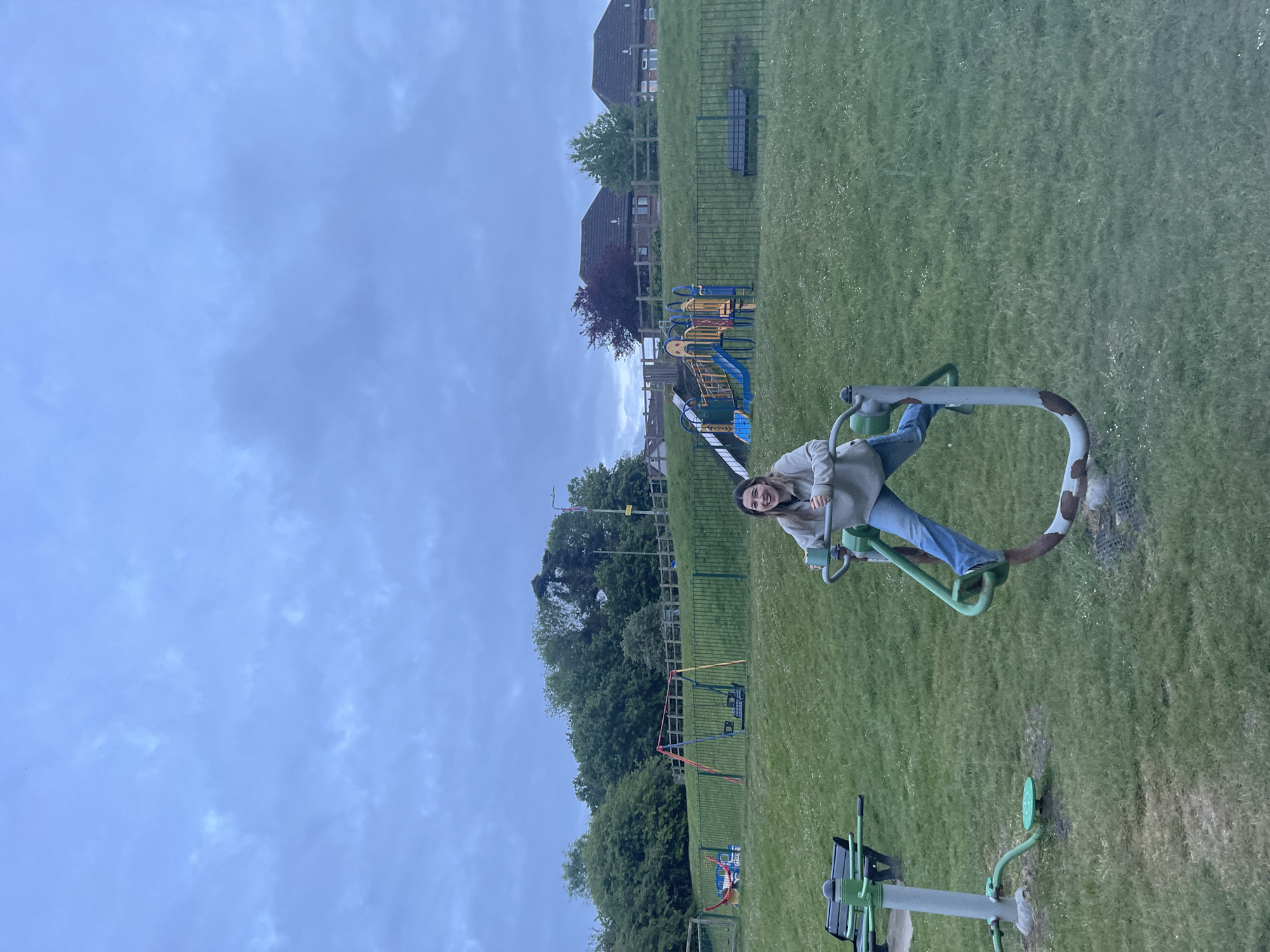 The image depicts a grassy park in Maidstone, United Kingdom, under an overcast sky. A young woman is sitting on a piece of outdoor gym equipment. Behind her, there is a playground with slides and swings. Further back, houses stand on a slight rise. The playground is enclosed by a green fence. Additional outdoor gym equipment is visible in the foreground. The grass is green and there are spots of white daisies.