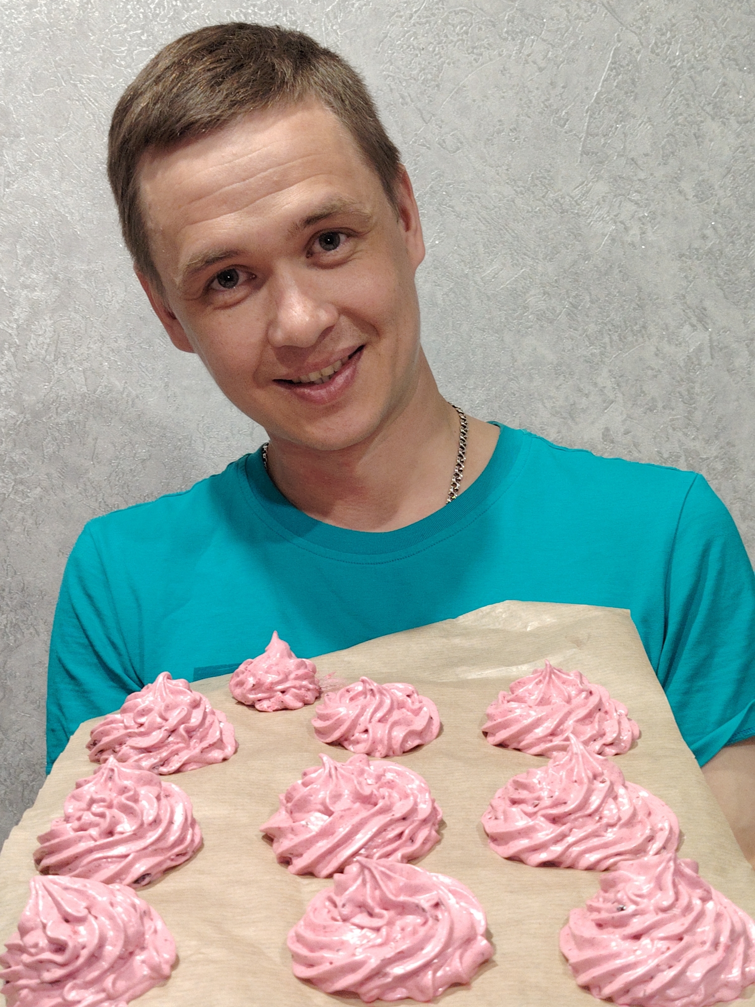 A smiling man with short brown hair and a silver chain necklace, wearing a turquoise t-shirt, is holding a tray of pink meringue-like desserts. The desserts are arranged on parchment paper. The background is a light gray, textured wall. The photograph appears to have been taken indoors in Chaikovsky, Russia.