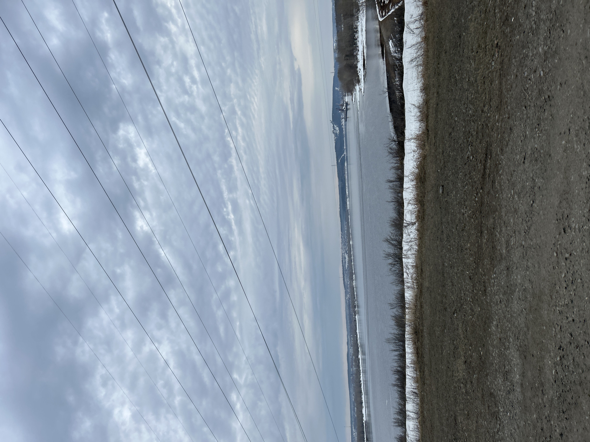 The image shows a view near the Kama River in Chaikovsky, Russia. Power lines dominate the left side of the frame against a cloudy sky. A body of water, likely the Kama River, runs diagonally through the center. The right side of the frame shows a shoreline with vegetation and a section of gravel or dirt road. The overall scene appears to be a somewhat desolate or rural area.