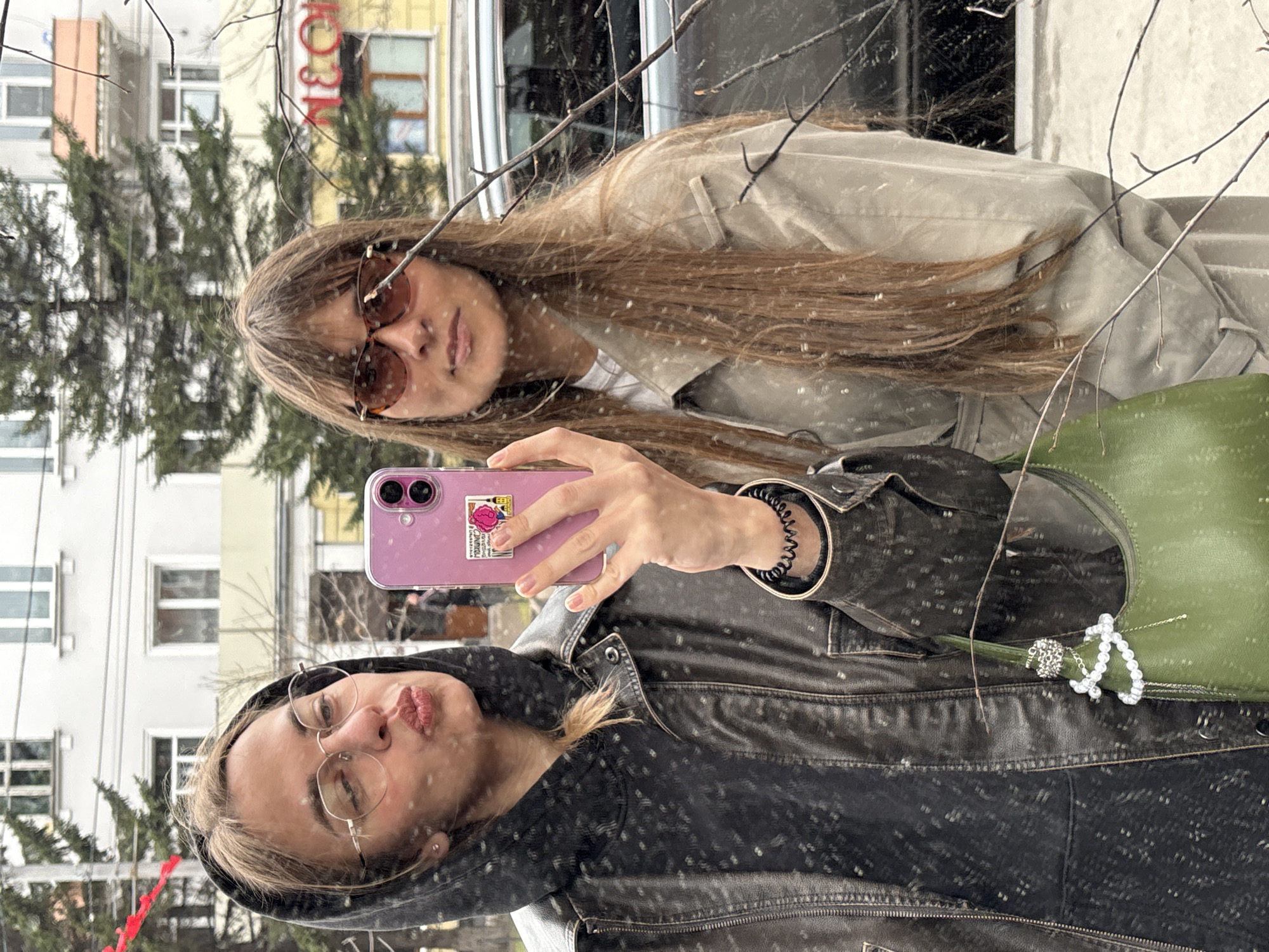 Two young women are reflected in a window. One is wearing a black jacket, the other a tan coat and brown sunglasses. The first woman holds a pink phone and is puckering her lips. Behind them can be seen a building with a sign in Cyrillic, a Christmas tree, and other buildings, reflecting elements of Irkutsk, Russia. It is snowing.