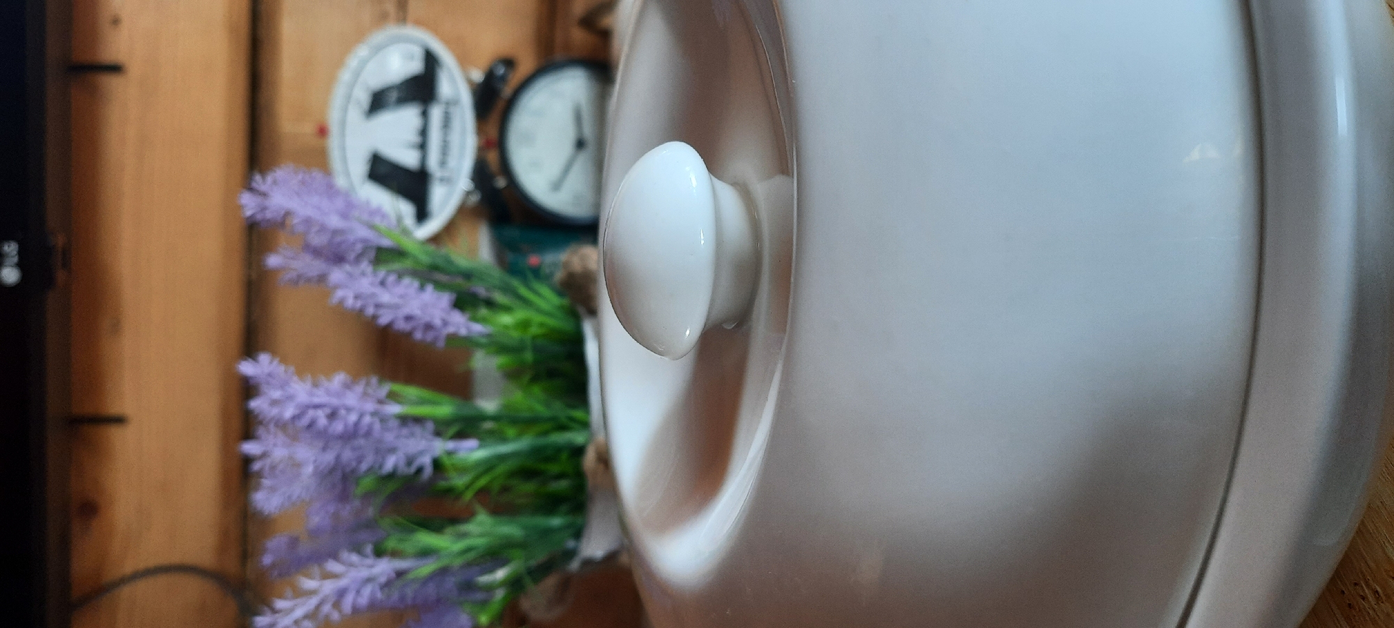 The image shows a close-up of a white lid with a round knob on top, part of a larger ceramic pot or dish. In the background, a blurred arrangement of artificial lavender flowers in a white pot is visible. Behind the flowers, a vintage-style mechanical scale with a white face and black hands is partially visible, and an oval decorative object with a silhouette of buildings is affixed to it. The backdrop appears to be made of wooden planks. The lighting suggests an indoor setting. The image does not provide any information about the city of Borogontsy, Russia, or any specific location.