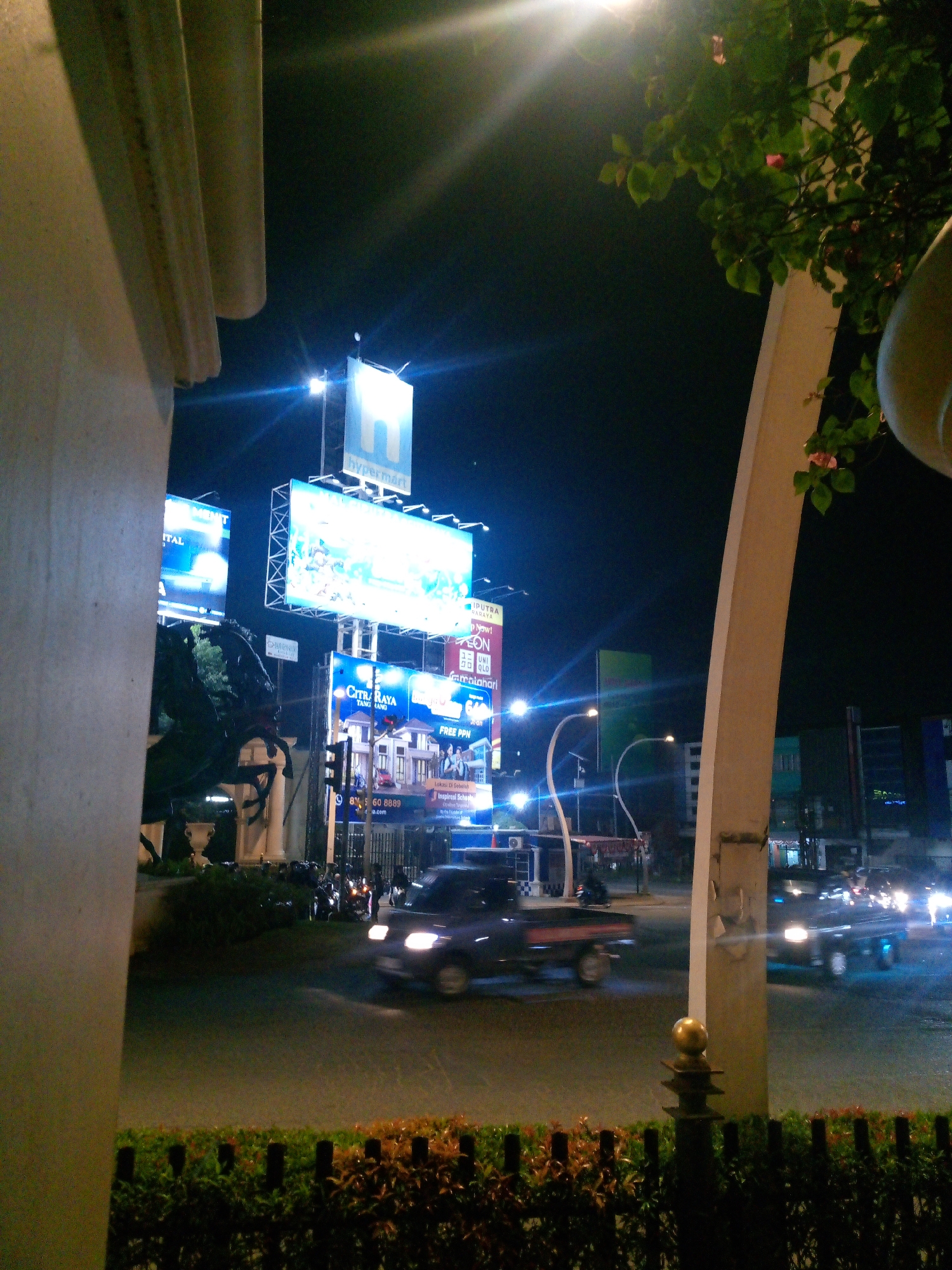 The image depicts a street scene at night in Bitung Jaya, Indonesia, characterized by illuminated billboards and traffic. A prominent "H hypermart" billboard is visible, along with other advertisements for "CYTRA RAYA" and "UNIQLO". Several vehicles, including cars and motorcycles, are present on the road, with headlights and taillights creating streaks of light due to motion blur. A dark-colored pickup truck is the most discernible vehicle in the foreground. Traffic lights are visible, indicating an intersection. The environment appears to be an urban setting with buildings in the background. A decorative fence with lush greenery lines the bottom of the frame, and a large white architectural element, possibly a pillar or archway, frames the left and right sides of the image. The overall impression is that of a busy commercial area at night.