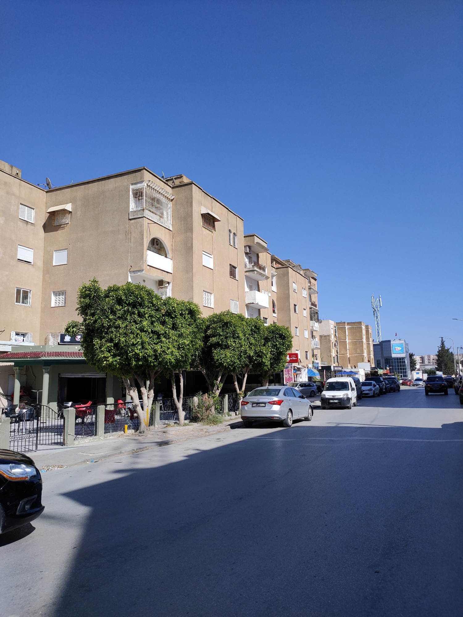 A street scene in Tunisia under a clear blue sky features a row of multi-story buildings on the left, characterized by their beige facade, white-trimmed windows, and balconies. Two lush green trees stand in front of the buildings, their branches full and dense. A cafe or restaurant with outdoor seating is visible behind a black metal fence. A black car is parked on the left side of the road in the foreground.
The street itself is paved and lined with parked cars, including a silver sedan, a white van, and several other vehicles. The buildings continue down the street on both sides, with varying architectural styles. In the distance, a cellular tower is visible. The overall impression is that of a moderately busy urban street on a sunny day.