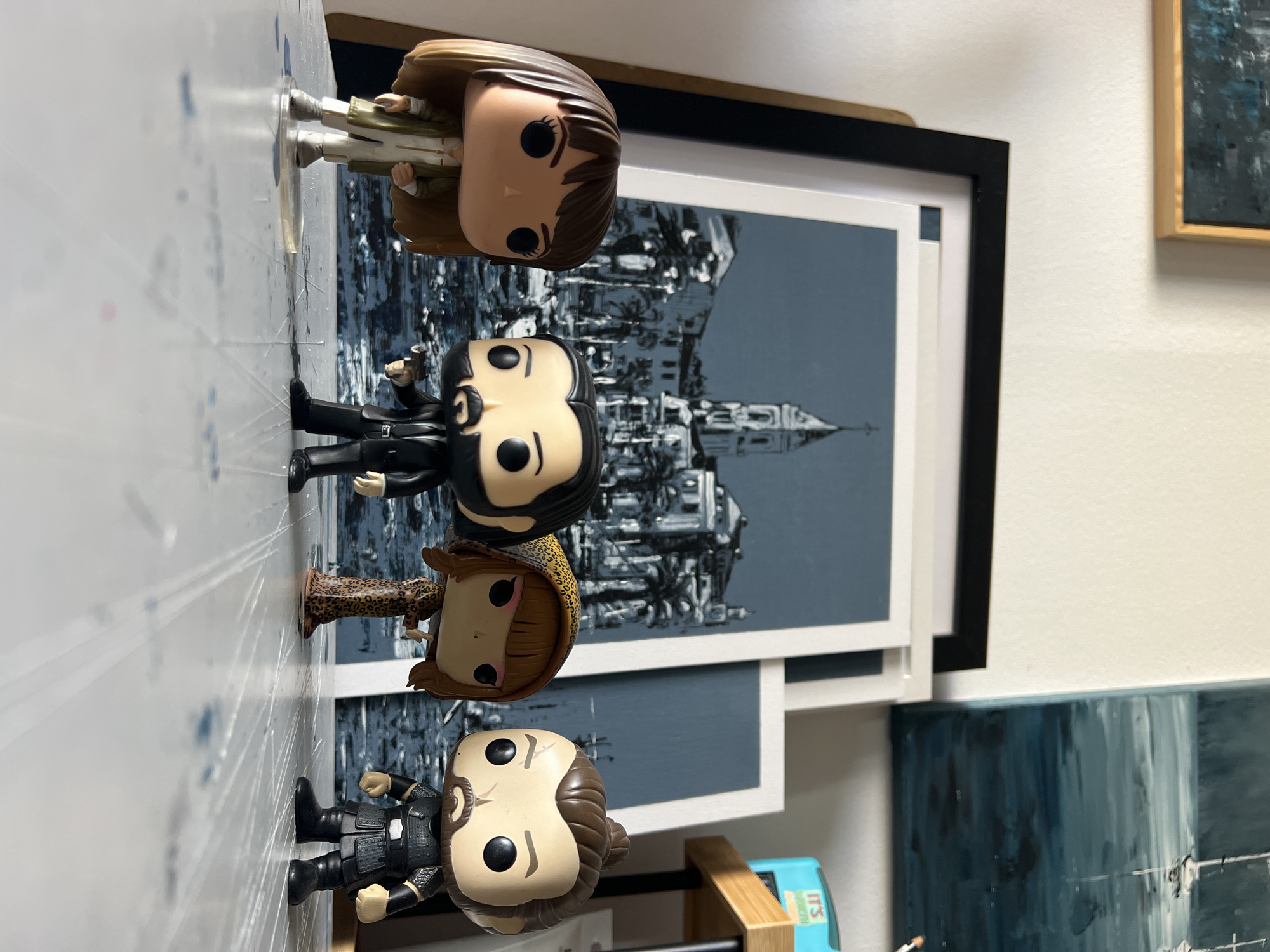 Four Funko Pop figures are displayed on a white surface, possibly a table, in front of several framed artworks. From left to right, the figures are a woman with brown hair and a green jacket, John Wick holding a gun, a woman in a leopard-print dress, and a man with dark hair tied up and wearing armor. The artworks behind them appear to be modern paintings, one depicting a city with a tower, and another with boats on water. The scene is lit by natural light, creating shadows on the white surface. The location is not Mollet del Vallès, Spain, but rather an indoor setting.