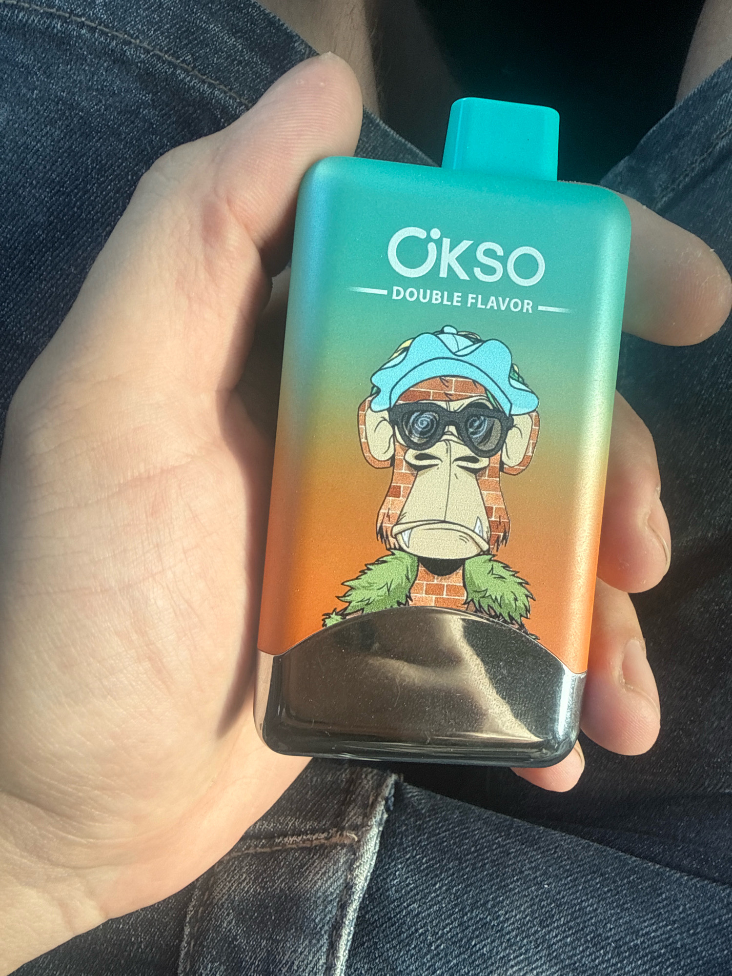 A hand holds a light blue and orange vape pen with a cartoon ape wearing sunglasses and a hat. The vape pen has the logo "CIKSO" and the text "DOUBLE FLAVOR". The background appears to be denim fabric. The image was taken in the city of Gorzów, Poland.