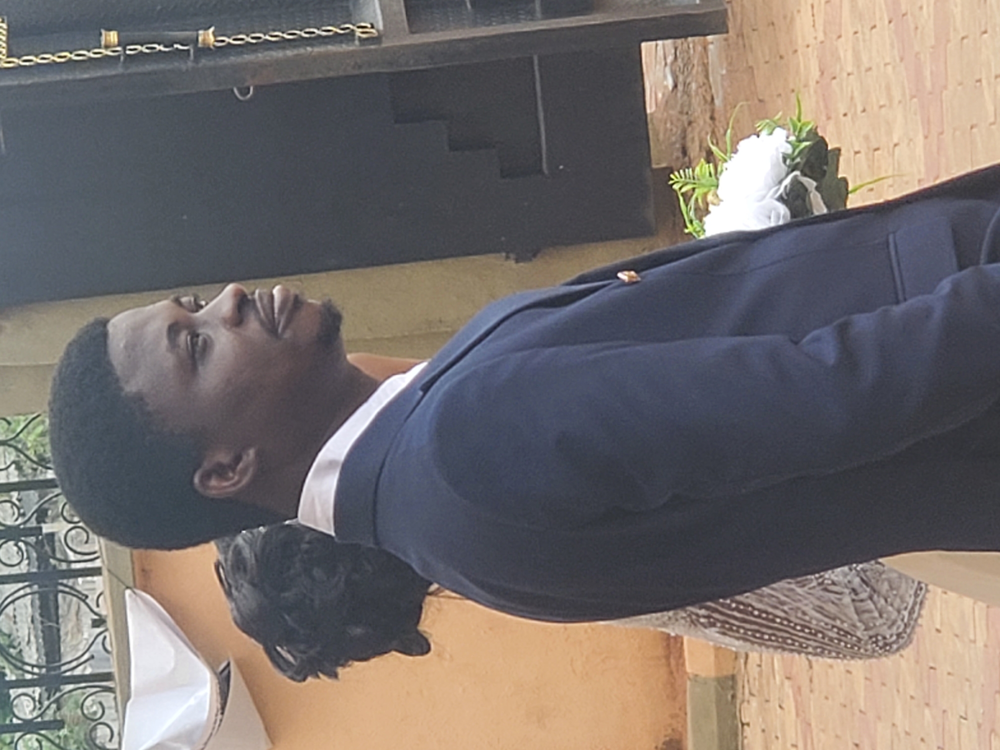 A man in a navy blue suit and white shirt is pictured outdoors. He has dark hair and a trimmed beard. The background suggests a building exterior with a decorative metal gate on the left and a tan wall. To the right, a white floral arrangement is visible. The location is suspected to be Onitsha, Nigeria.