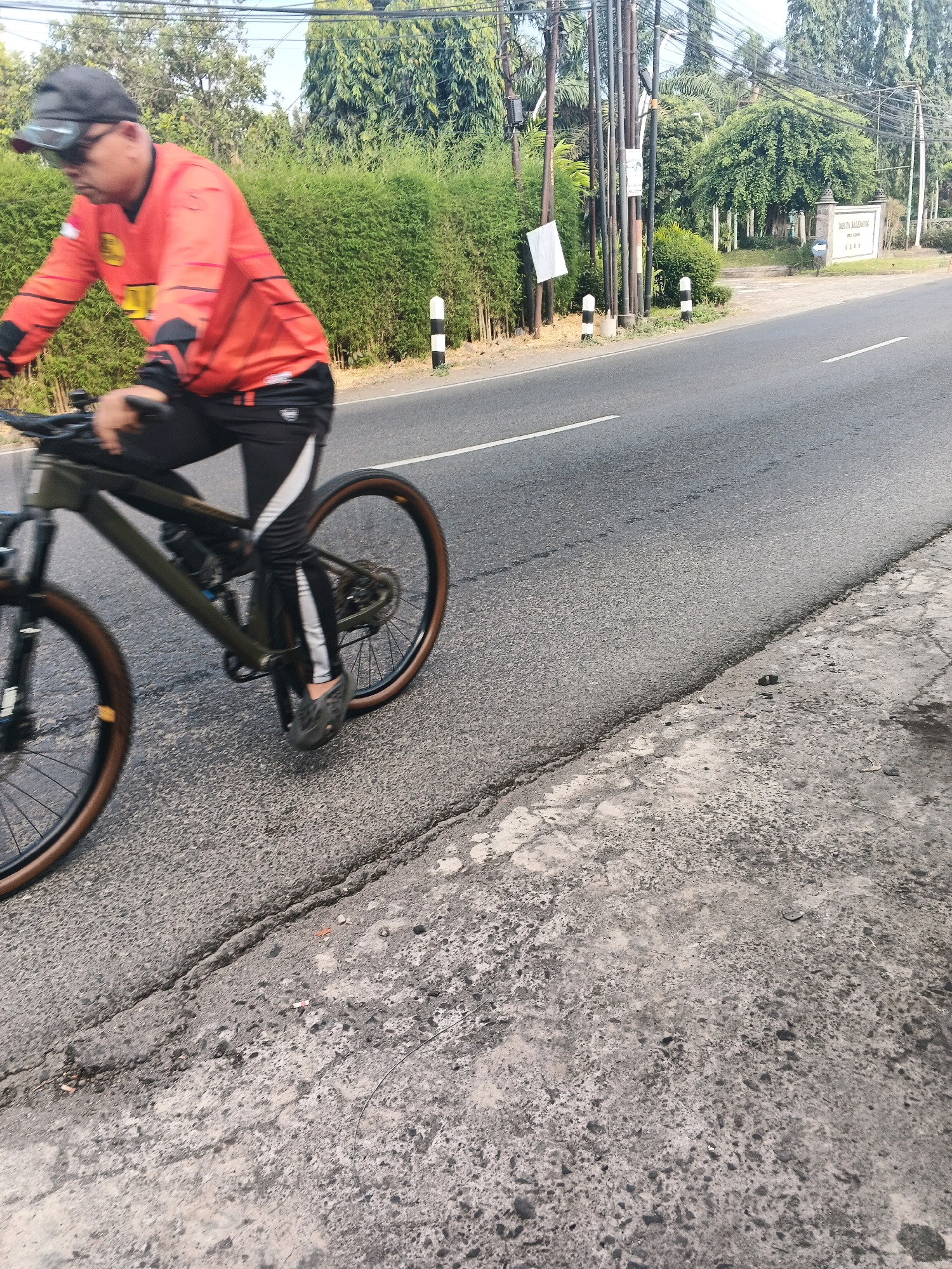 The image depicts a person riding a bicycle on a paved road in Ungaran, Indonesia. The cyclist is wearing an orange long-sleeved jersey with black accents and black cycling pants with white stripes. They are also wearing a black cap and sunglasses. The bicycle appears to be a mountain bike with a dark frame and brown tires. The road is asphalt with a white line marking the lane. To the left of the road, there is a lush green hedge, followed by a line of bamboo and utility poles. In the background, there are trees and a sign for "MELAN BALEMONK". The ground on the right side of the road is a cracked concrete surface. The scene is captured in daylight with soft shadows.