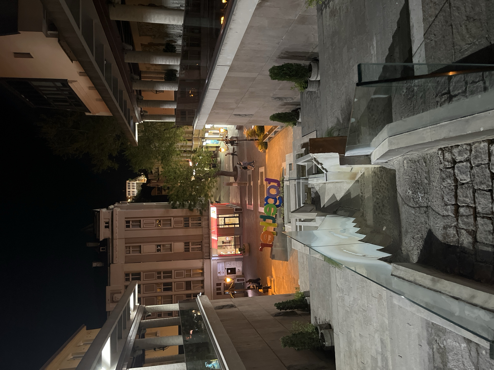 This is a nighttime shot in Plovdiv, Bulgaria, looking down a pedestrian street. Buildings flank the street, illuminated by street lights and shopfronts. People are strolling along the street. Colorful letters spell out "Together." A glass barrier is in the foreground. The pavement is light-colored stone. The sky is dark.