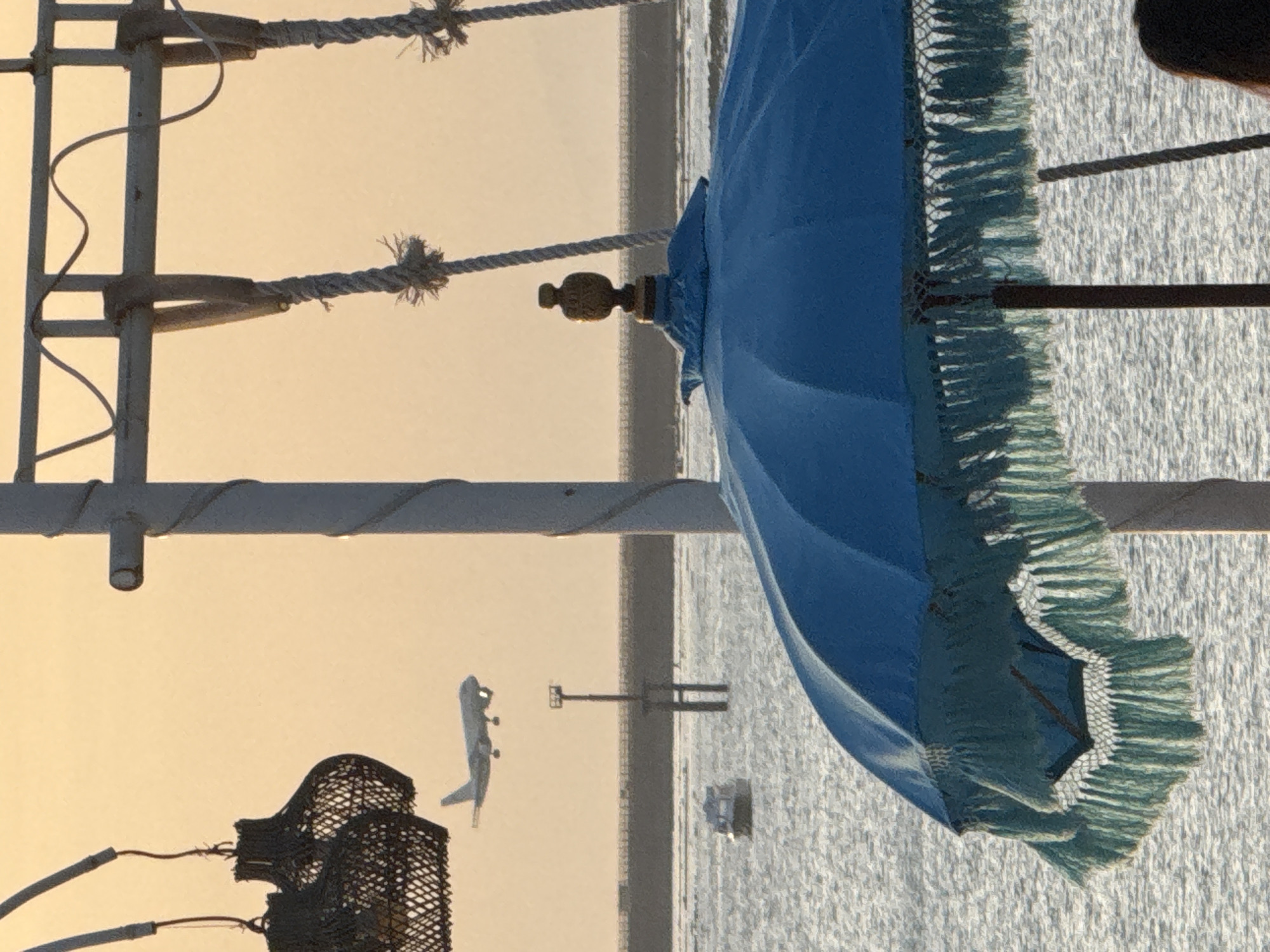 This is a photograph taken at Kuta, Indonesia. A blue-fringed umbrella is present in the foreground. A rope-rigged structure is visible to the left of the umbrella, and an airplane takes off in the distance. The background is a light gradient suggestive of sunset or sunrise over the water. There's a fishing basket hanging on the structure.