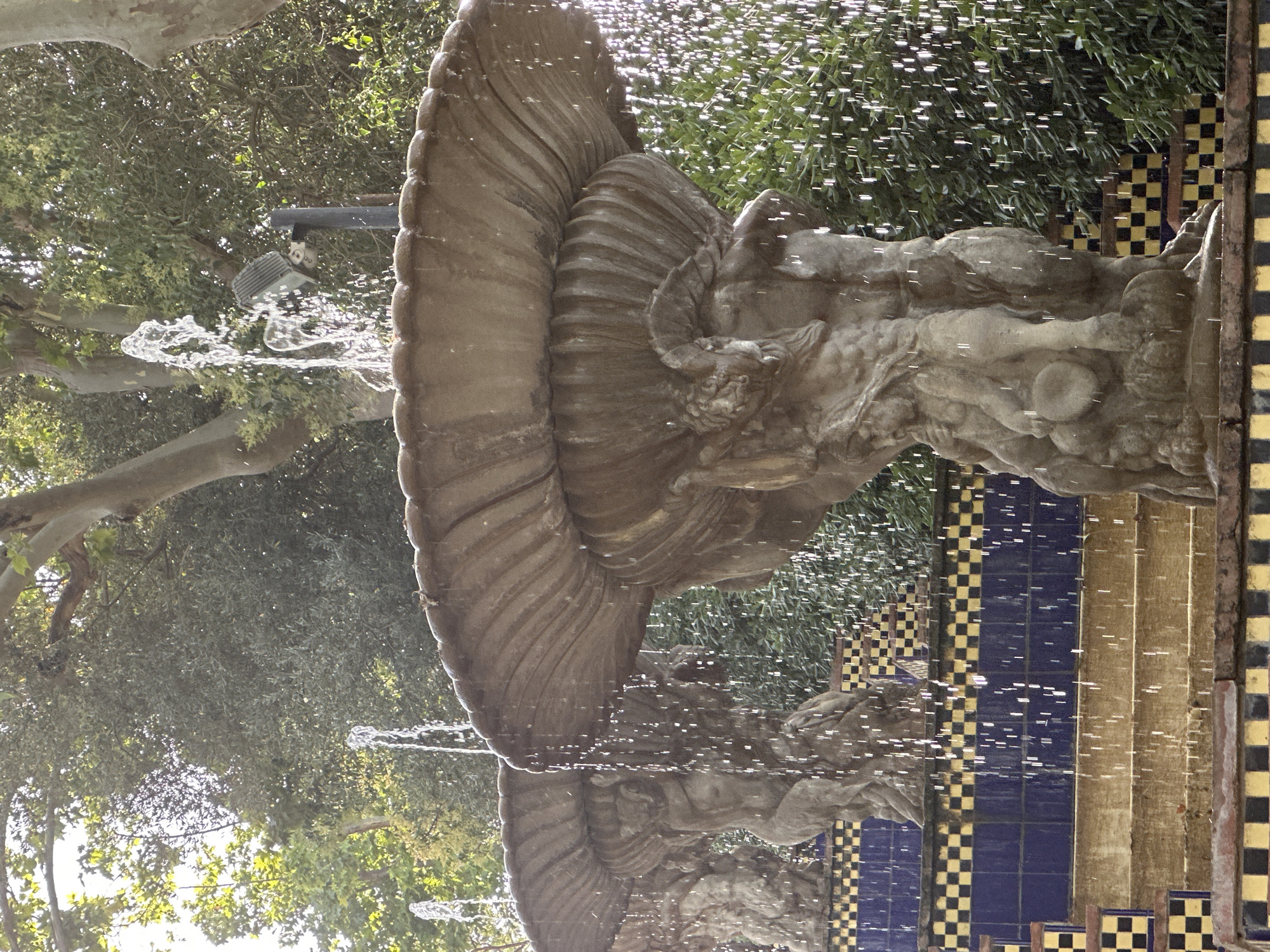 The image depicts a stone fountain with multiple tiers, carved with figures that appear to be Atlantes or similar mythical beings supporting the upper basins. Water is actively flowing from the fountain, creating a cascade of droplets that are illuminated by sunlight. The fountain is situated on a tiered staircase decorated with blue and yellow checkered tiles. Lush green foliage forms the background, suggesting an outdoor setting. In the distance, trees with visible branches and leaves are present, and the sky is partially visible through the canopy. A modern spotlight is visible mounted on a pole behind one of the water jets. The scene appears to be a detail shot of a larger architectural or garden feature, possibly within a public park or historical site. The architecture and tiling suggest a location in Barcelona, Spain.