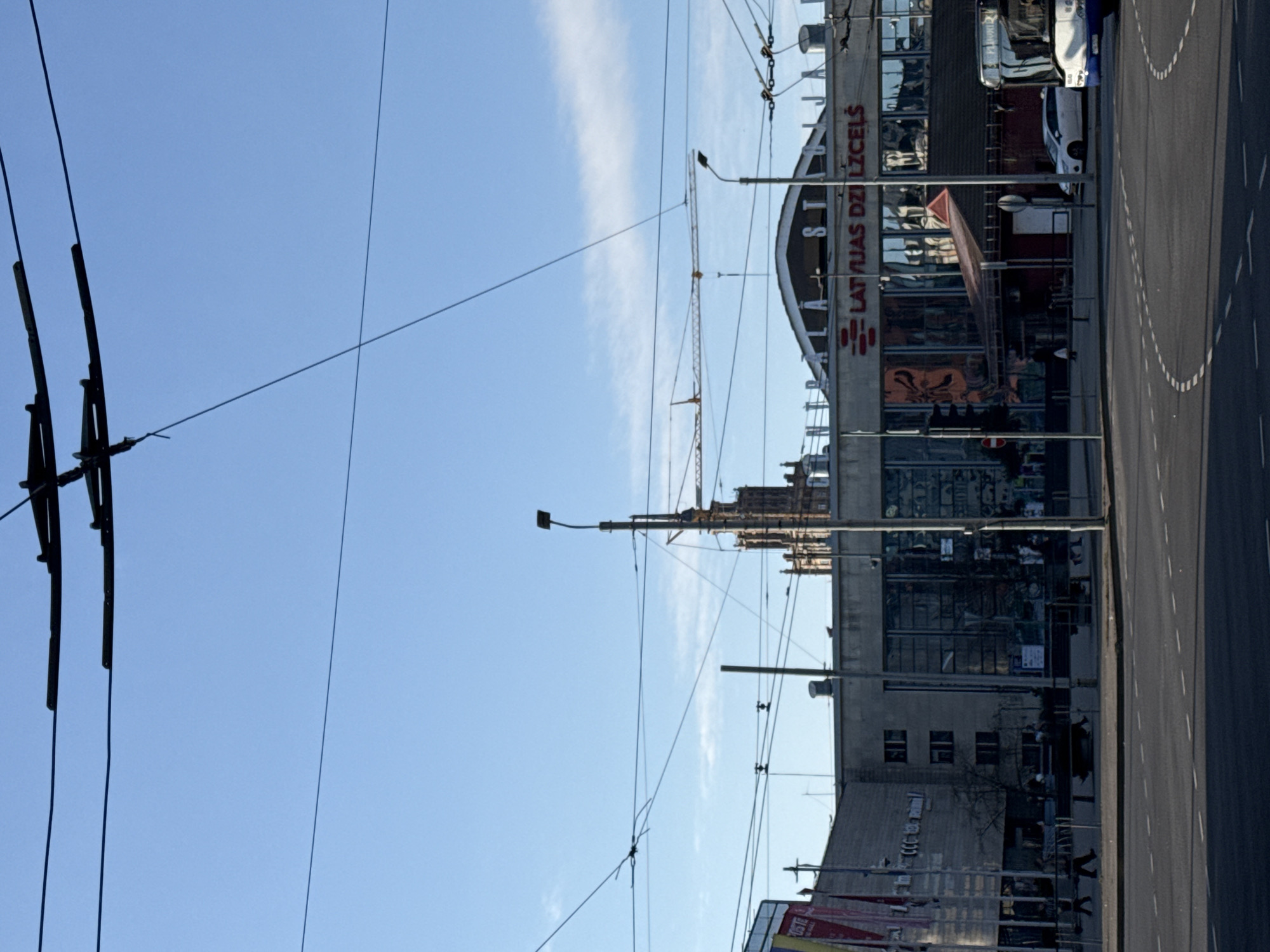 The image shows a street scene in Riga, Latvia, featuring the "Latvijas Dzelzceļš" (Latvian Railway) building. The building has a distinct arched roofline and prominent signage. A construction crane is visible near the building. An overhead tram line runs along the street. There are cars parked next to the building. The sky is blue with some clouds. A road runs alongside the building with painted markings on the surface. The view is slightly tilted.