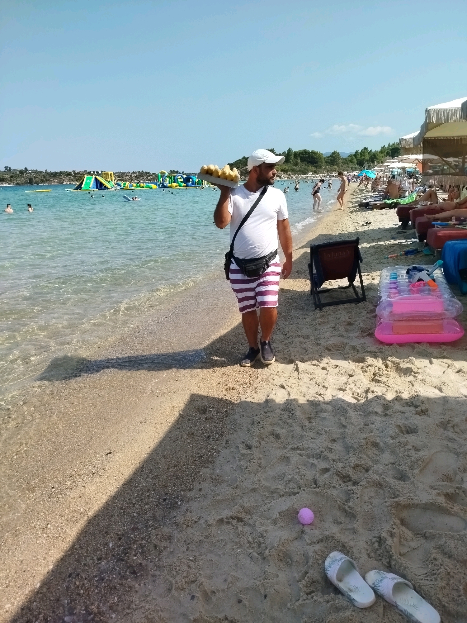 A man wearing a white t-shirt, striped shorts, and a white cap is walking on a sandy beach in Sithonia Municipal Unit, Greece. He is carrying a tray of what appears to be food. The beach is crowded with people swimming in the clear, shallow ocean and sunbathing on lounge chairs. In the background, there's a large inflatable water park in the sea. The sky is clear and blue.