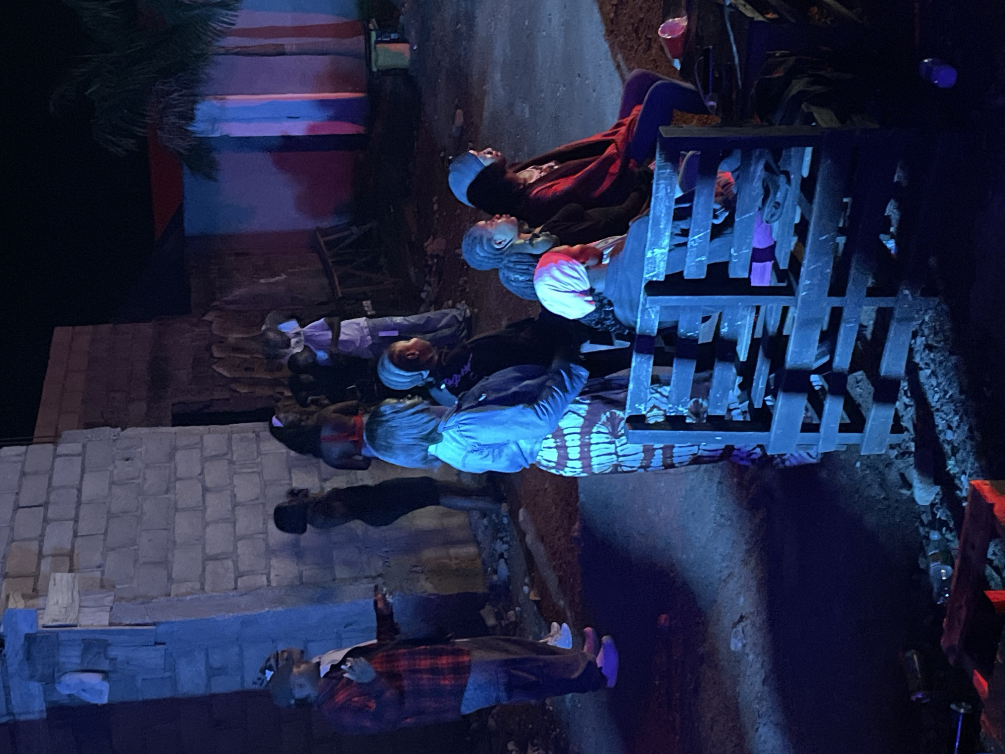 In Durumi, Nigeria, at night, a group of people are gathered outdoors, illuminated by artificial lights, casting long shadows. The scene appears to be a street or an open area with unfinished brick buildings and a palm tree visible. Several individuals are standing and sitting, some appearing to watch something or engage in conversation. One person in the foreground, wearing a red and black plaid shirt, is standing and gesturing. A group of women are seated on a wooden structure, facing away from the camera. The lighting suggests a festive or communal gathering.
