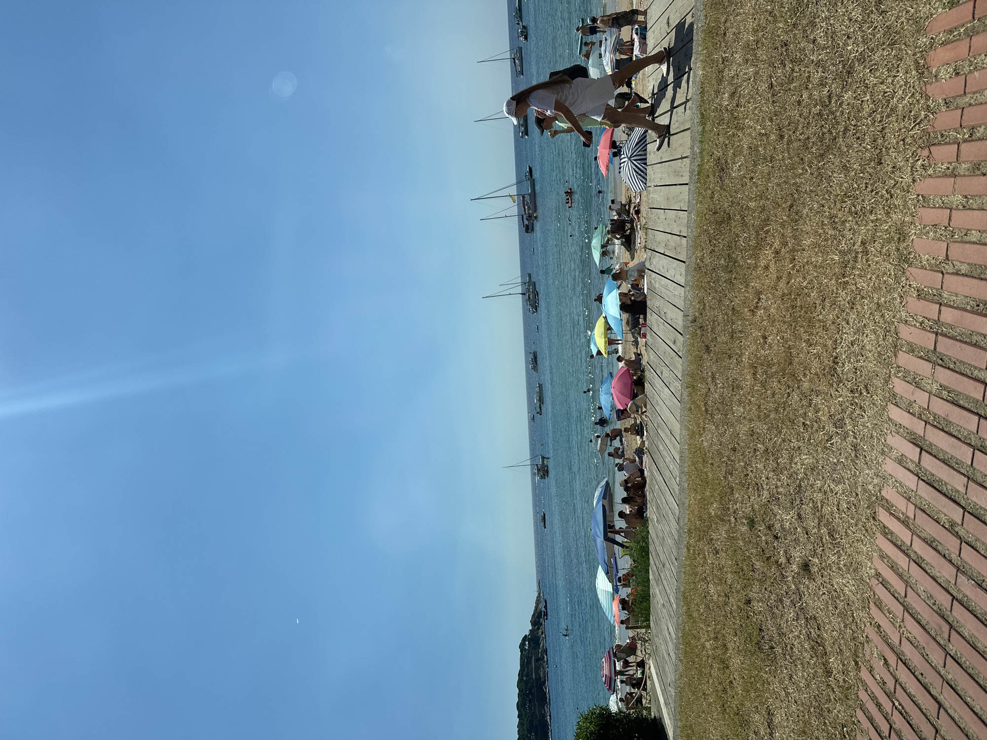 A wide-angle shot captures a sun-drenched beach scene in Sant Feliu de Guíxols, Spain. The foreground features a wooden walkway and a patch of dry grass, leading to a bustling beach filled with people. Colorful umbrellas dot the sandy shore, providing shade for beachgoers who are sitting, standing, and walking. The sea stretches out beyond the beach, dotted with numerous sailboats and smaller vessels. In the distance, a lush green headland juts out into the water. The sky is a clear, bright blue with a hint of lens flare in the upper right corner. Two women are walking on the wooden walkway in the middle ground, one wearing a white cap and the other a black cap.