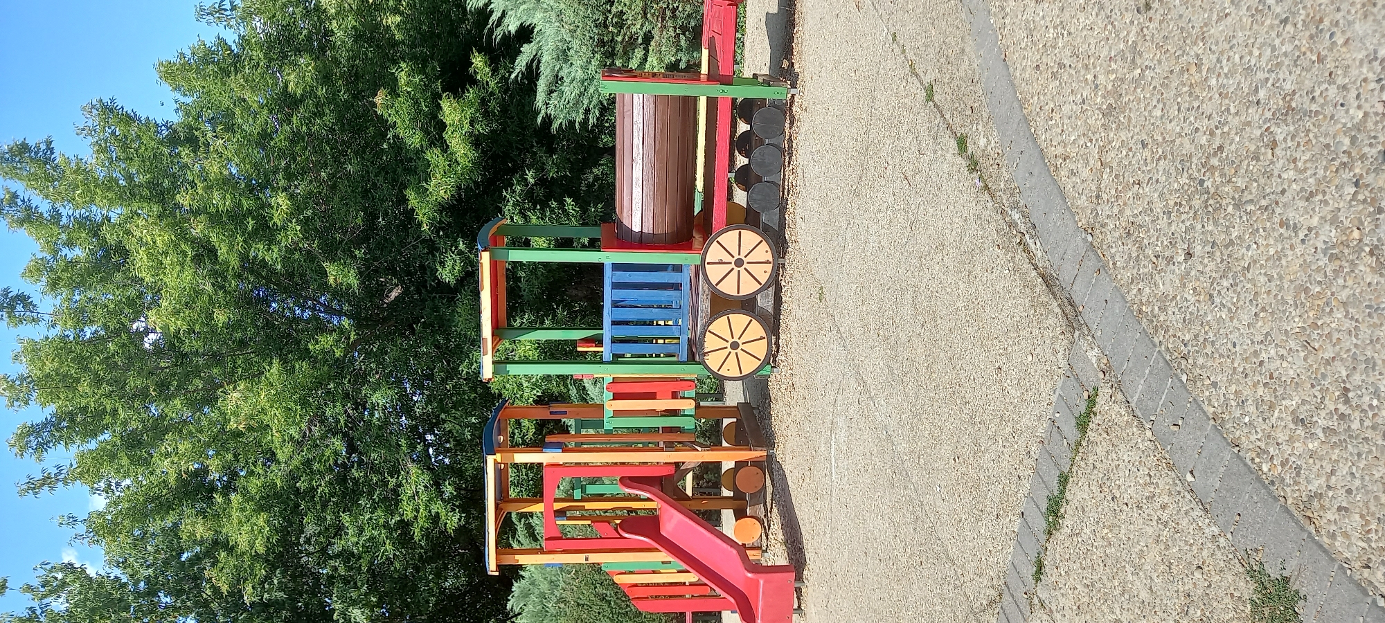 A vibrant, multi-colored children's playground structure resembling a train is situated on a sandy ground with concrete pathways in the city of Silistra, Bulgaria. The playground features a red slide, blue railings, wooden wheels with yellow accents, and a brown cylindrical section. The structure is framed by lush green trees and bushes under a clear blue sky. There are no people visible in the image.