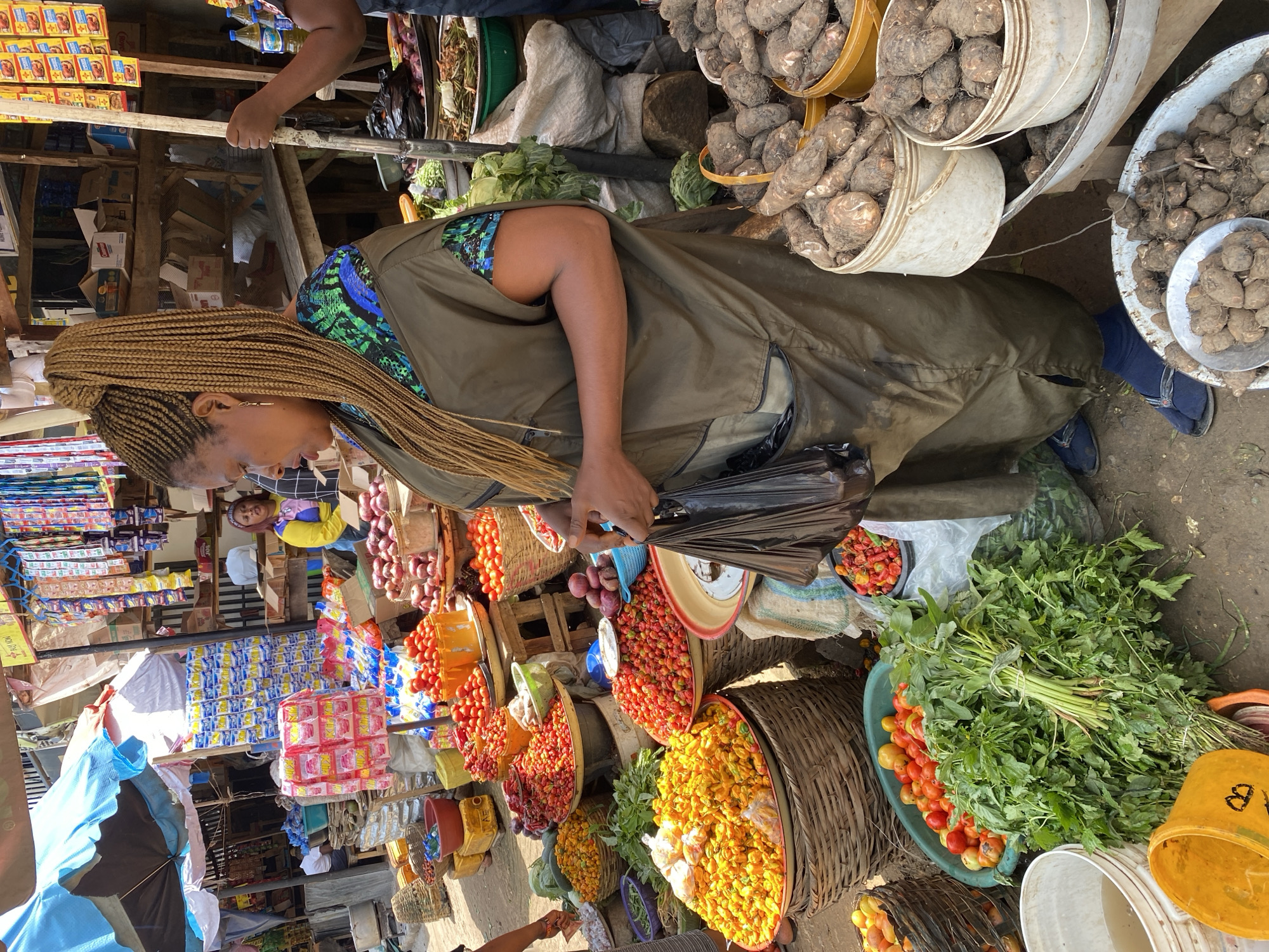 The image depicts a bustling market scene in Jos, Nigeria. The primary subject is a woman with long, blonde braided hair wearing a green vest over a colorful patterned top and dark pants. She is partially obscured by a large green garment and appears to be handling black plastic bags.
The setting is an outdoor market with stalls filled with various produce and goods. Prominently displayed are large quantities of tubers, possibly yams, in buckets and a metal basin. Other visible produce includes tomatoes, chilies, onions, and leafy green vegetables. Various baskets are used to hold and display these items. In the background, shelves are stacked with boxes of packaged goods, some with colorful labels. A blue tarp is visible on the left side of the image, and several plastic containers, including yellow buckets, are scattered throughout the scene.
The image appears to have been taken during the day, with bright lighting suggesting it's not nighttime. The overall atmosphere seems active and busy, typical of a marketplace. There are no explicit emotional cues visible from the main subject, but the vibrant colors of the produce and the sheer volume of goods imply a lively commercial environment.
There is some visible text on the yellow bucket in the lower right corner, which appears to be the letter "B" in yellow. The context of Jos, Nigeria, suggests a West African market environment.