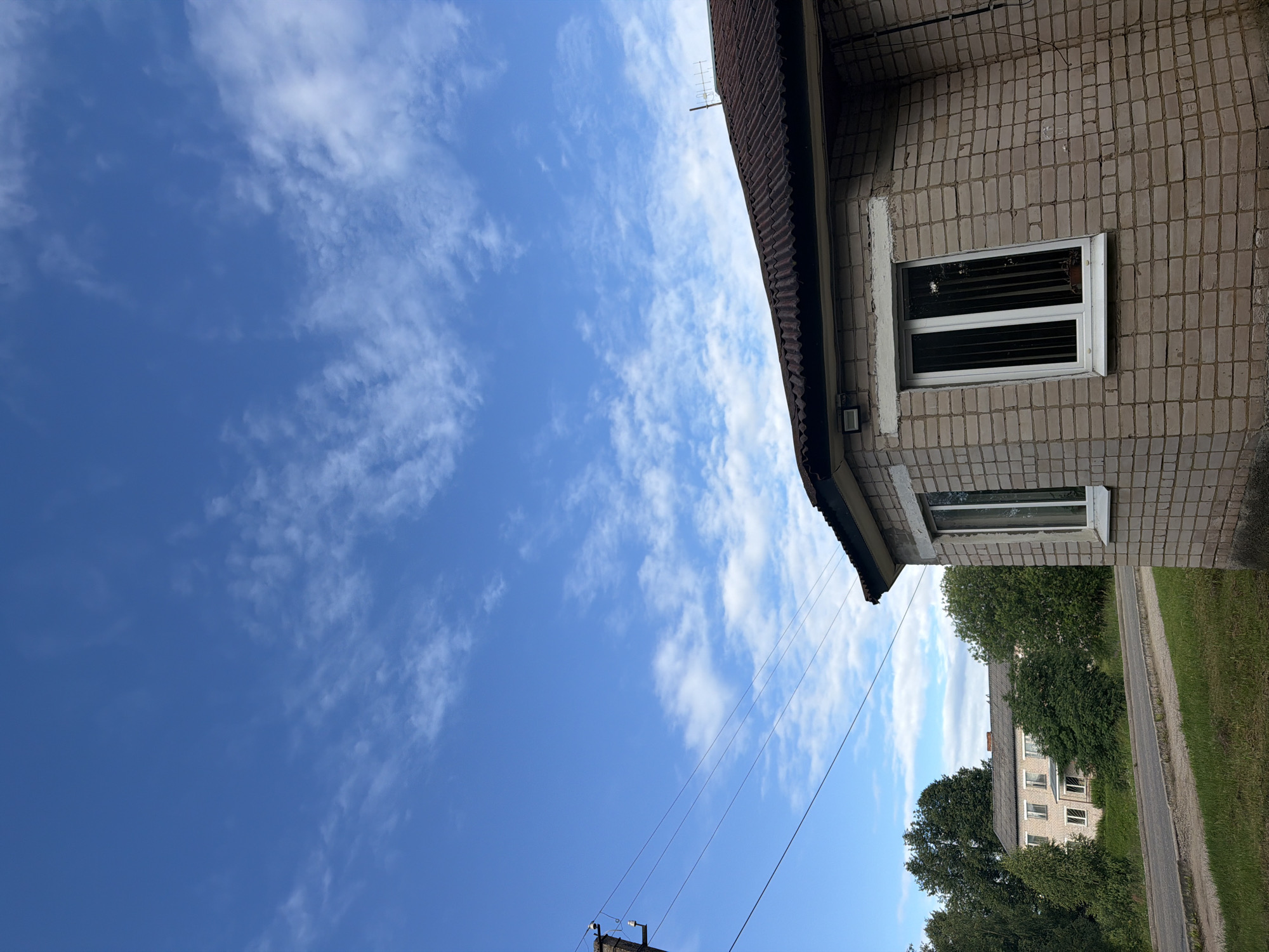 The media file depicts an exterior view of a building and the sky. The building appears to be constructed with light-colored brick and has a dark-tiled roof. A white-framed window with horizontal blinds is visible on the brick wall. Electrical wires crisscross the frame, connecting to a utility pole in the lower portion of the image. In the background, a street runs alongside a grassy area, with more buildings and lush green trees lining the road. The sky is a vibrant blue, populated with scattered white, wispy clouds, suggesting fair weather. The lighting indicates it is daytime, likely late morning or afternoon, given the clear sky and the brightness of the scene. There is no discernible text in the image. The overall impression is one of a quiet, residential area under a bright, cloud-filled sky. The scene is captured from a slightly low angle, looking up towards the building and sky. The orientation of the image is such that the sky occupies the left half and the building and its surroundings the right half, suggesting it may have been taken in a vertical or slightly tilted orientation.