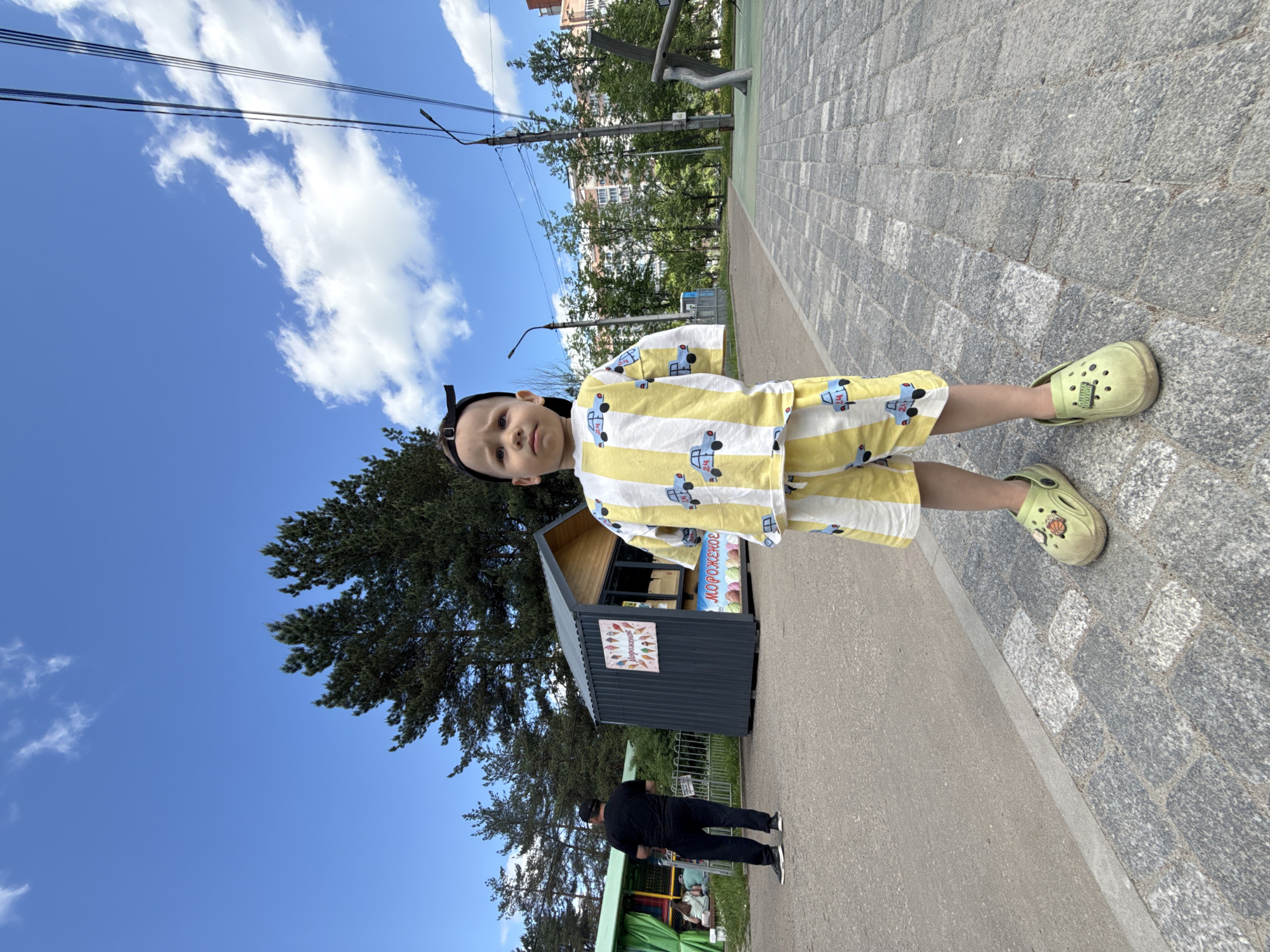 The image depicts a young child in a yellow and white striped pajama set with car designs, wearing light green clogs, standing on a paved sidewalk. Behind the child is a small, dark-colored ice cream stand. A man in black clothing is walking near a green structure behind the ice cream stand. A tree is to the left of the ice cream stand, and the sky with clouds is visible above. The location is Rusanovo, Russia.