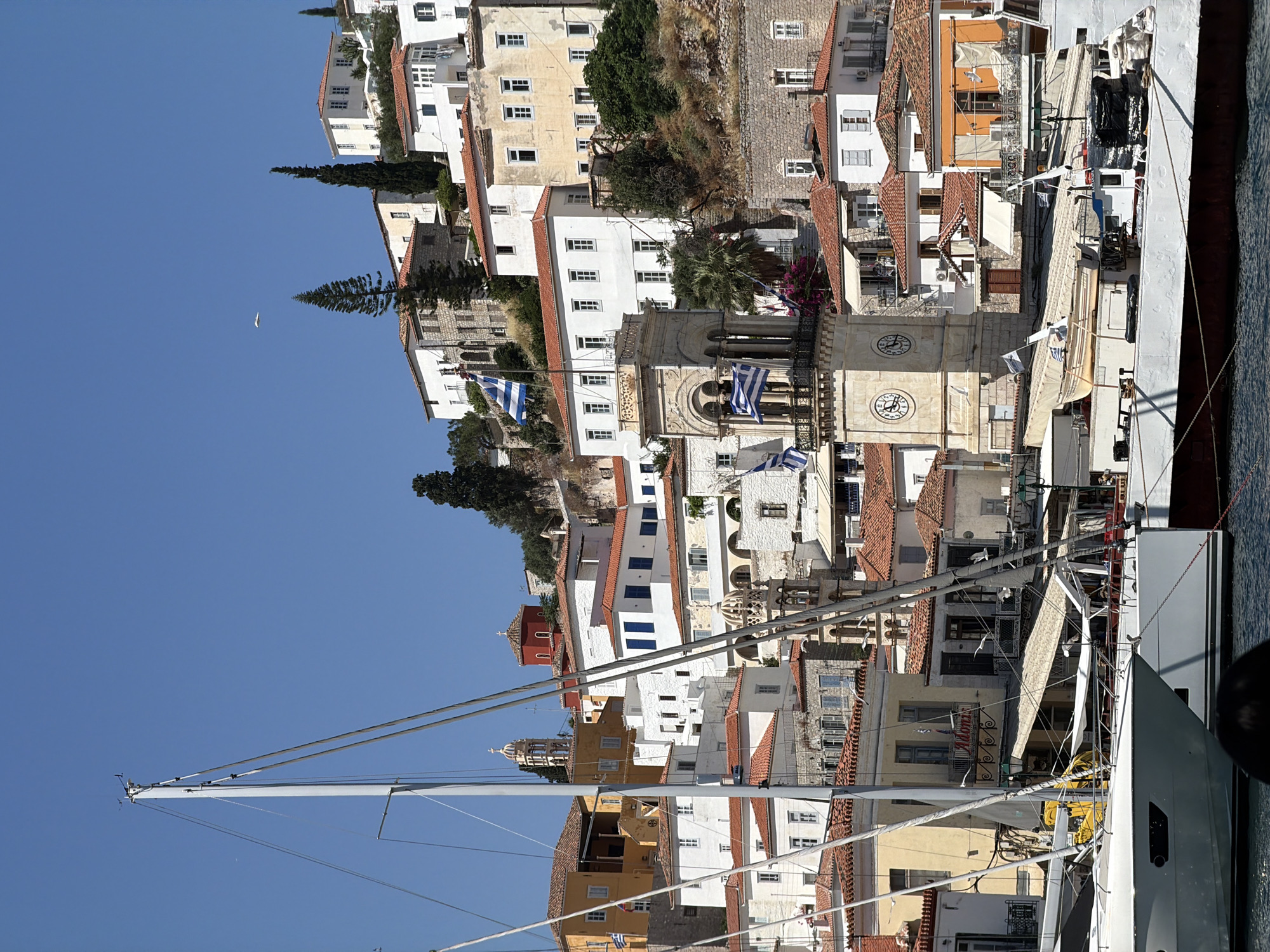 The image depicts a vertical shot of Hydra, Greece, a port city in the Greek Islands. The cityscape consists of buildings clustered together, mostly in white, with red-tiled roofs. The Greek flag is visible on the buildings. A boat is positioned in front of the city, with its mast extending into the sky. The sky is clear and blue, and a bird can be seen flying. The architectural details of the buildings are highlighted. The image captures the coastal scenery of the island.