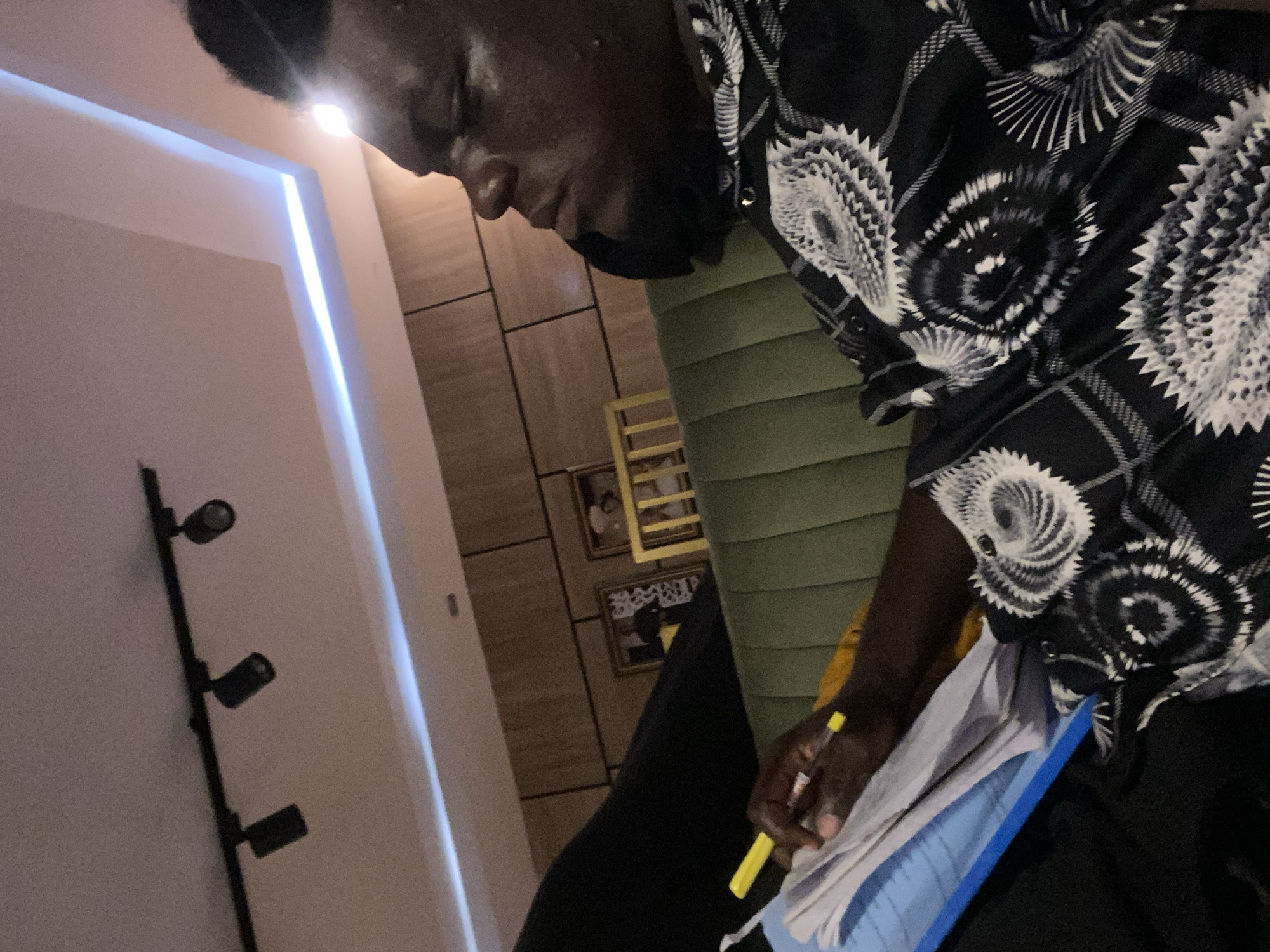 An indoor scene captured from a low angle features a dark-skinned person engrossed in writing on a stack of papers with a yellow pen. The person is wearing a black and white patterned shirt, and the papers are resting on a blue surface. The background includes a wall with decorative vertical grooves and framed photographs, along with a green ribbed surface, possibly part of a piece of furniture. A track lighting fixture with three spotlights is mounted on the left wall, and a vertical strip of light provides additional illumination.