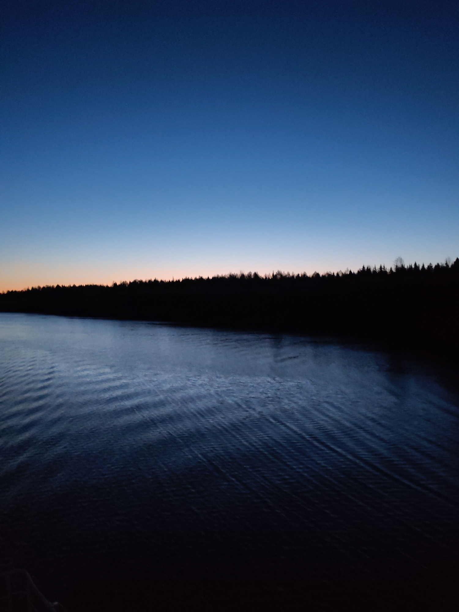 The image depicts a body of water, likely a lake or river, under a twilight sky. The sky transitions from a deep blue at the top to a faint orange glow on the horizon. Silhouetted trees line the distant shore. The water's surface shows subtle ripples and disturbances, suggesting a slight breeze or current. The scene is dimly lit overall. It is not possible to definitively locate the scene in the city of Uzhla, Russia, or ascertain any details about people or actions from the image alone.