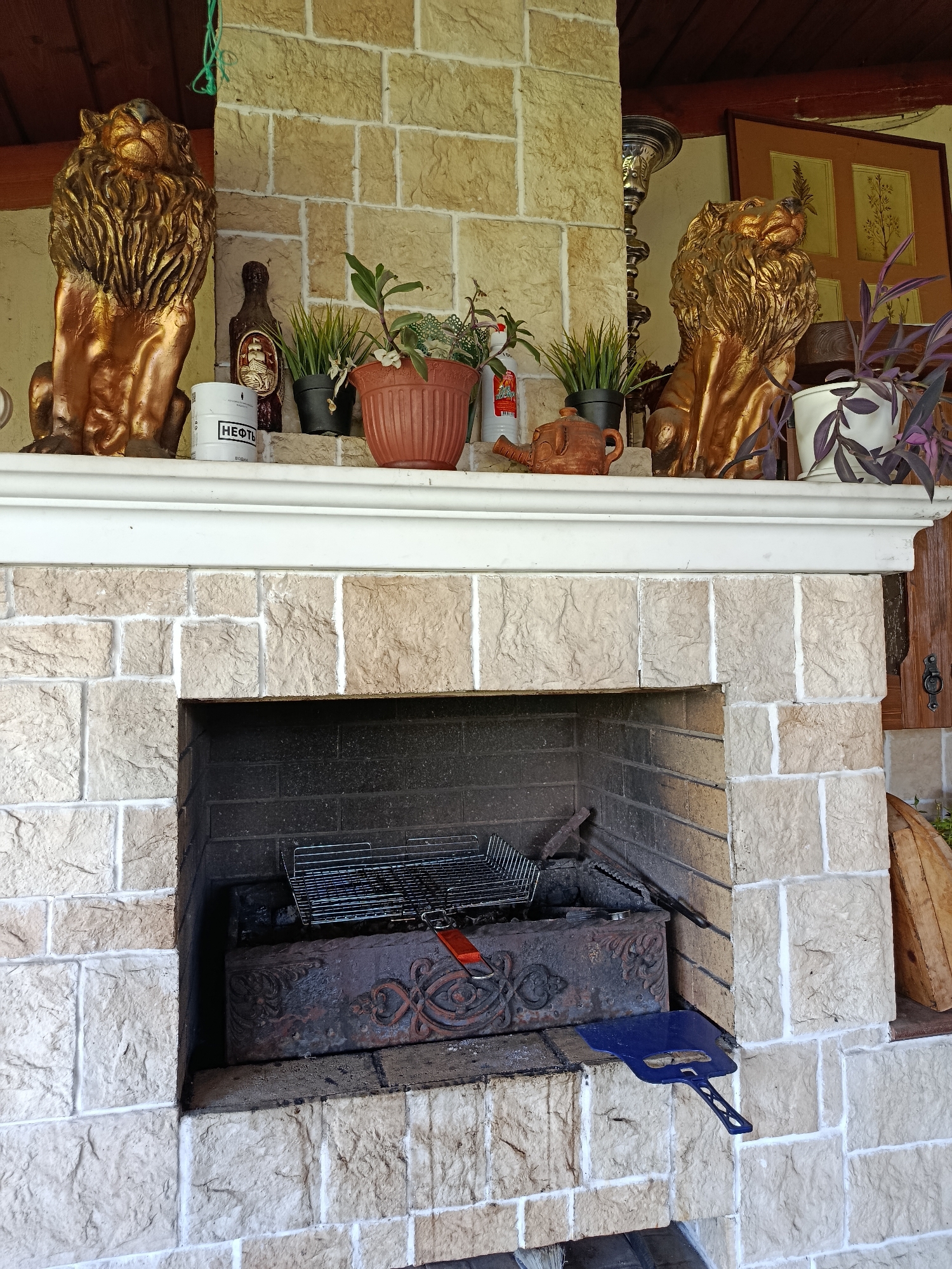 This is a medium shot, eye-level, outdoor photograph of a stone fireplace in Misaylovo, Russia. The fireplace has a light-colored stone facade and a dark brick interior. A metal grill rack is placed inside the fireplace. On the mantelpiece above the fireplace are two gold lion statues, several potted plants, a decorative teapot, a bottle, and a white can with Russian text. To the right of the fireplace, a wooden door is visible. A blue shovel rests on the stone facade to the right of the fireplace opening. The ceiling above the fireplace appears to be made of dark wood. The scene is well-lit.