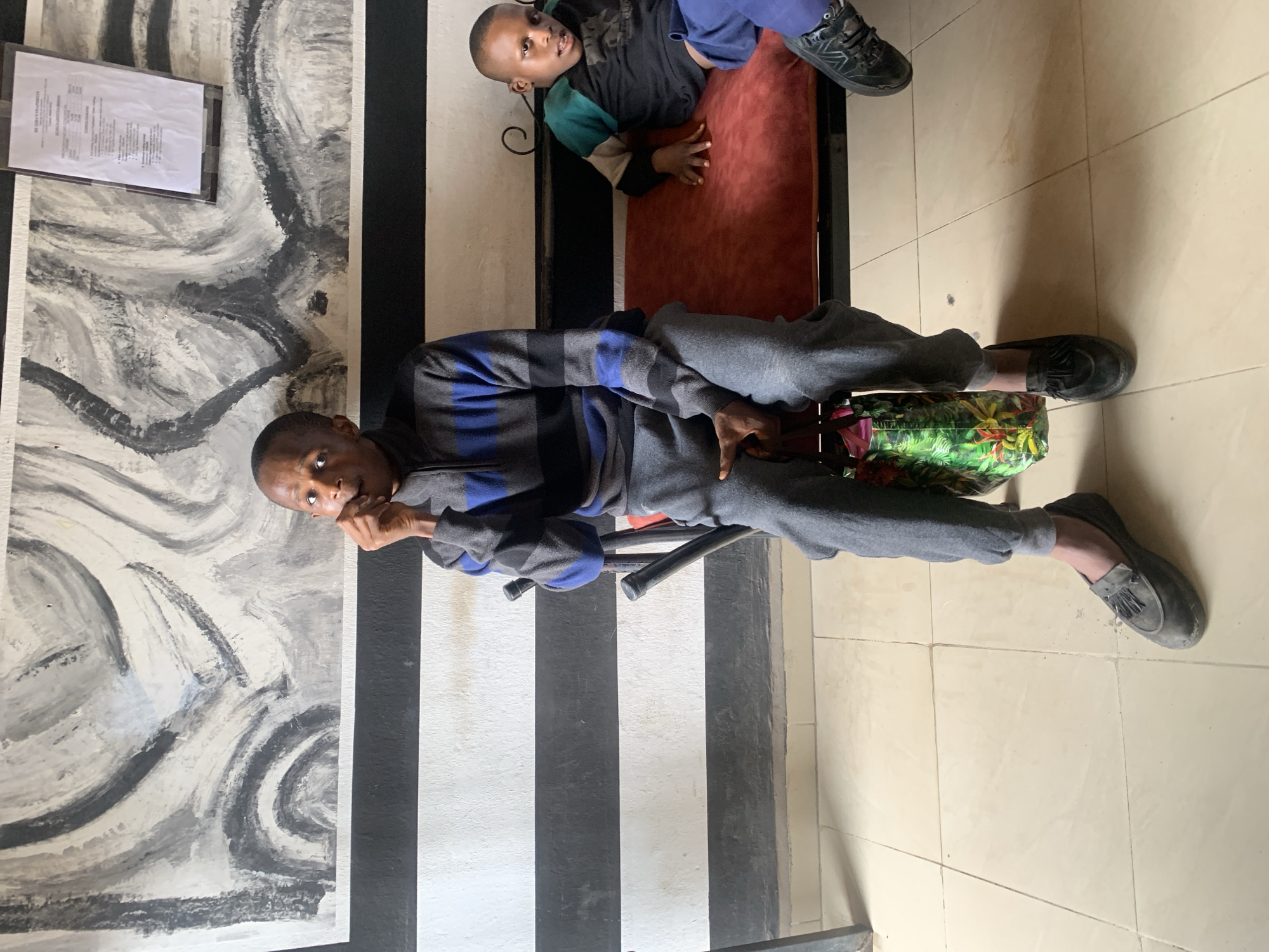 This indoor photograph, taken in Jos, Nigeria, features two males: an adult and a child. The adult, positioned closer to the viewer, is seated on a chair, looking directly at the camera. He wears a grey and blue striped long-sleeved top, grey sweatpants, and dark grey loafers with tassels. His right hand is near his mouth. Next to his feet rests a vibrant green bag adorned with a red and yellow floral or leaf pattern.
Behind the adult, a child is lying on a red cushioned bench, looking slightly to the side with an engaged expression. The child is dressed in a multi-colored long-sleeved shirt (black, green, and cream sections), blue pants, and dark sneakers.
The background wall features a distinct design: a section with bold black and white vertical stripes, and an adjacent section displaying an abstract mural in shades of grey, black, and white, characterized by swirling patterns. A framed document, containing text too small to be legible, is mounted on the mural section of the wall. The floor is covered with light-colored, possibly beige, tiles. The scene suggests a relaxed indoor setting.