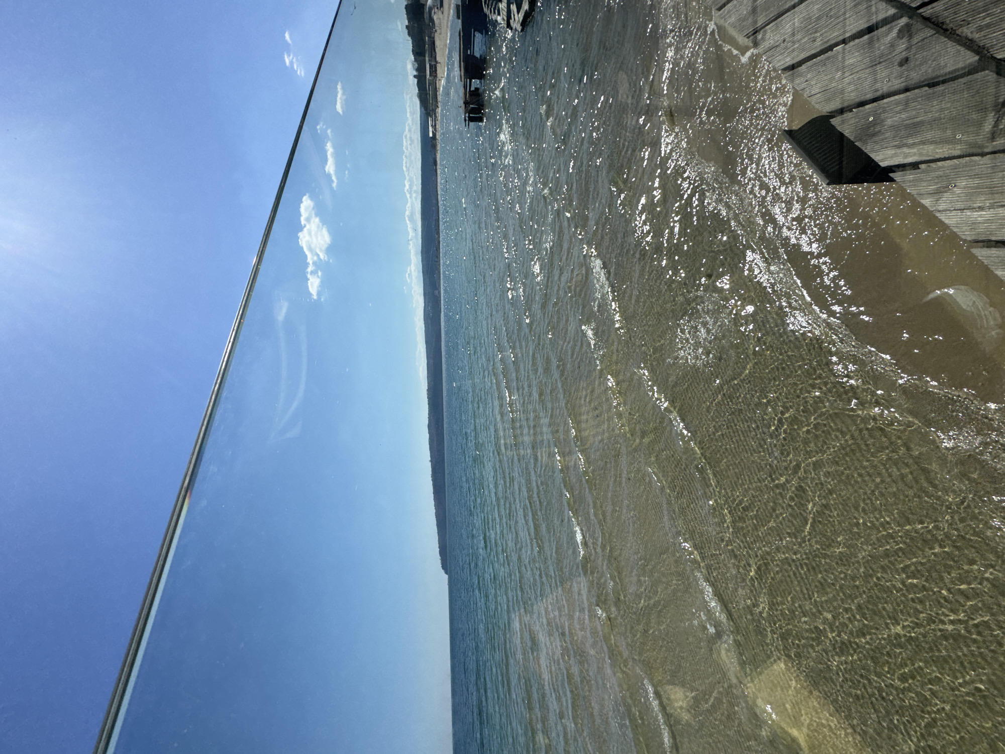 The image shows a body of water next to a wooden dock, with the sky visible on the left. The water appears to be shallow and clear, with visible ripples and a sandy bottom. The sky is blue with a few white clouds. The wooden dock is on the right, with planks running horizontally. The image appears to be taken from a low angle, with the water filling most of the frame. The location is likely in Varna, Bulgaria, given the presence of a beach and the Black Sea. There is no presence of people.