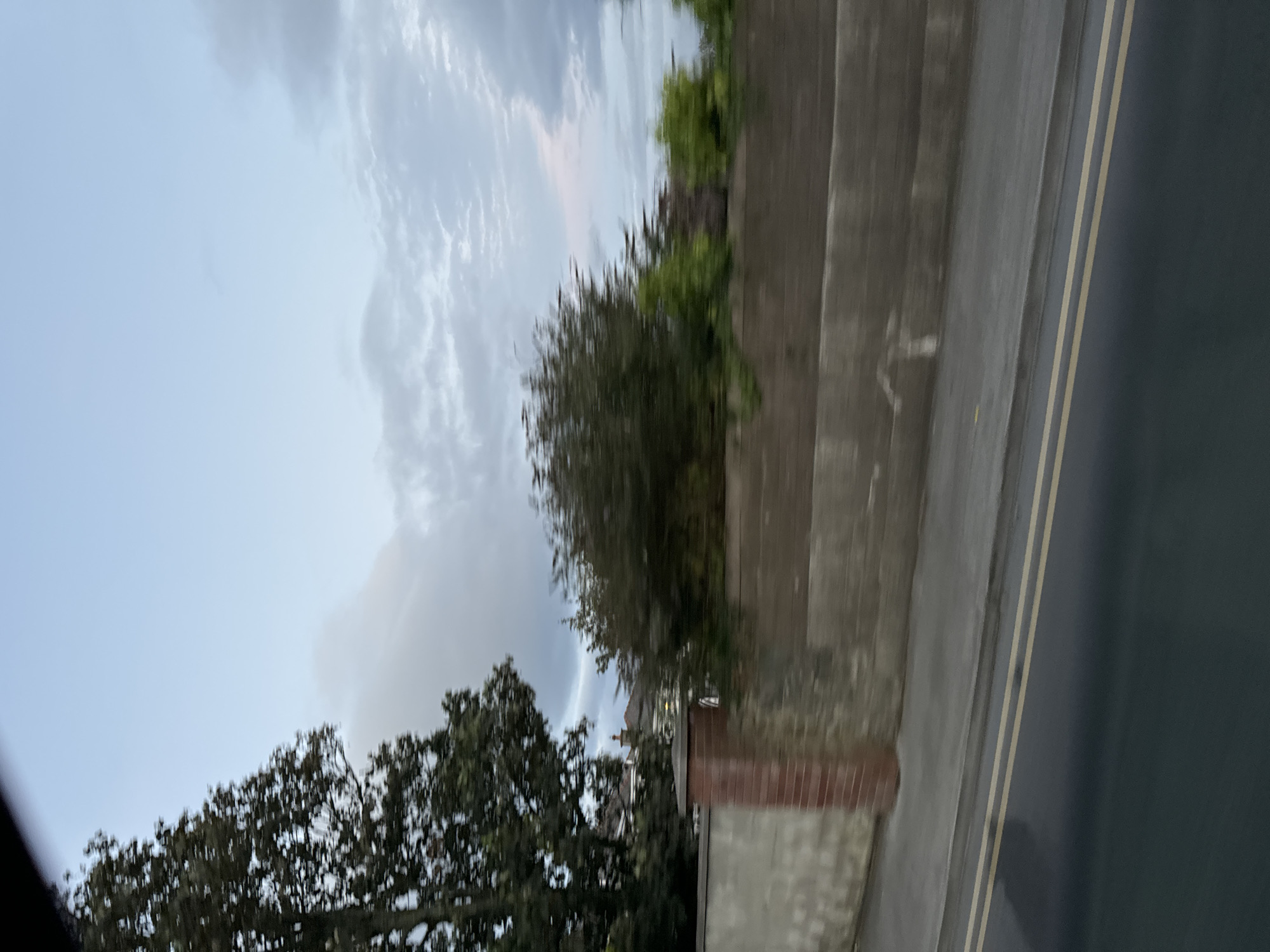 The image shows a motion-blurred view of a residential street in Tramore, Ireland. A grey concrete wall with a brick pillar runs along the side of the road. Behind the wall, there are trees and glimpses of houses. The sky is overcast with clouds. The road in the foreground has double yellow lines, indicating a no-parking zone. The overall impression is of a suburban environment captured from a moving vehicle.