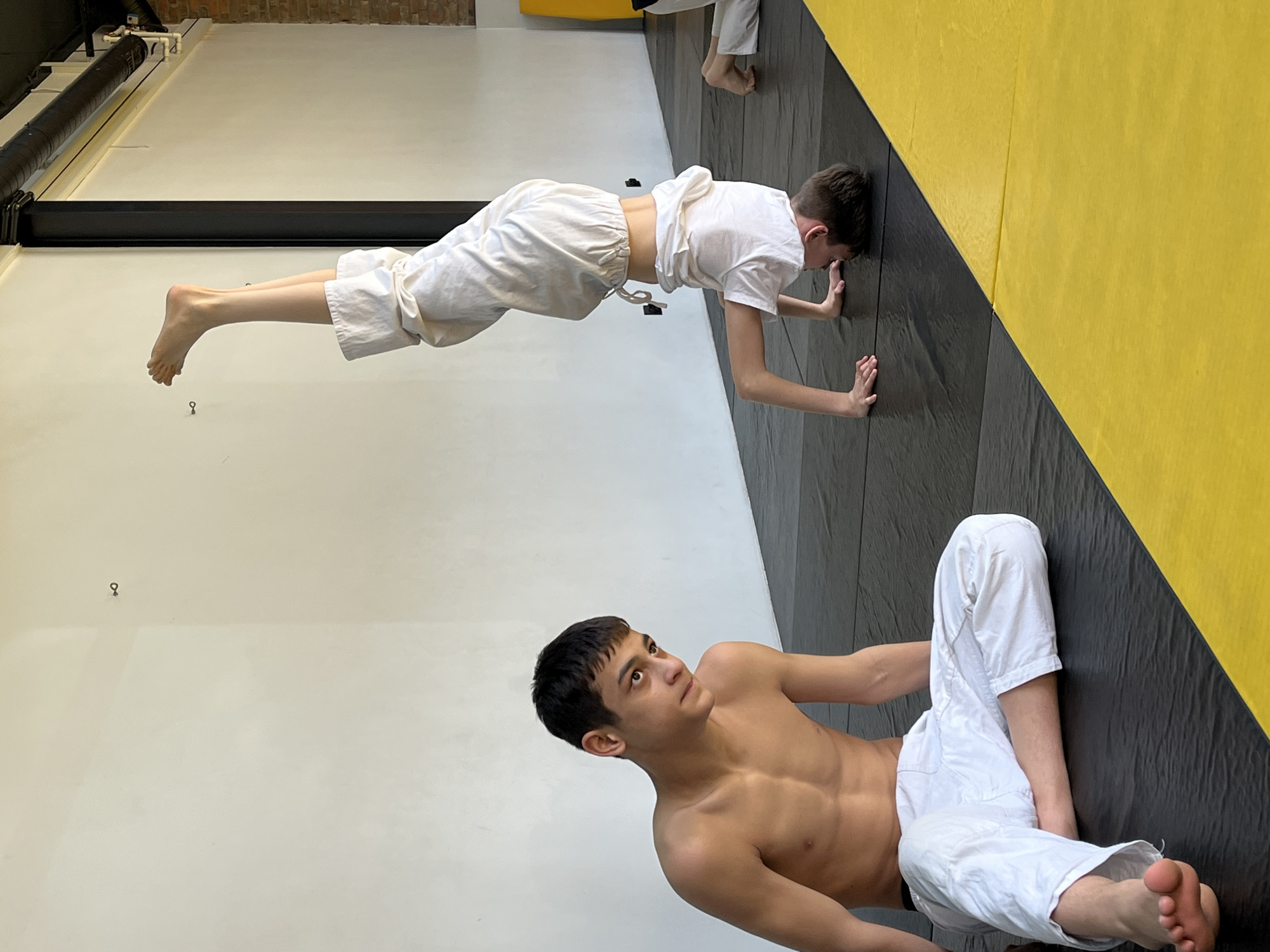 The image shows two males in what appears to be a gym or training space. One male, shirtless and wearing white pants, is lying on the floor. The other male, wearing a white t-shirt and white pants, is performing wall push-ups against a padded wall that is black and yellow. The location is potentially in Krasnoyarsk, Russia, but without additional visual or textual cues, this can not be confirmed.