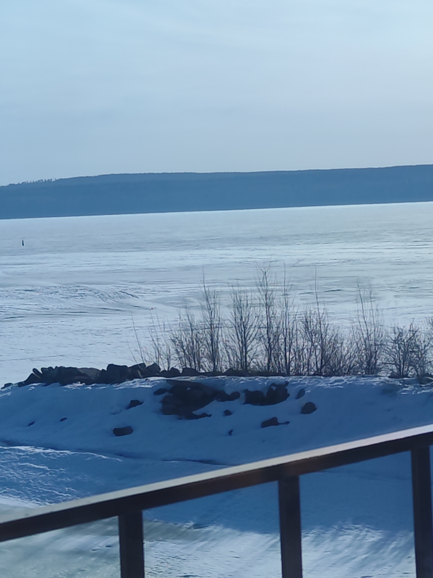 The image shows a frozen body of water, likely the Kama River, as Chaikovsky is located on its banks. In the distance, a tree-covered shoreline is visible. In the midground, there is a snow-covered bank with exposed rocks and bare trees. The foreground includes a portion of a metal railing. There is a structure in the water (the left side of the photograph).