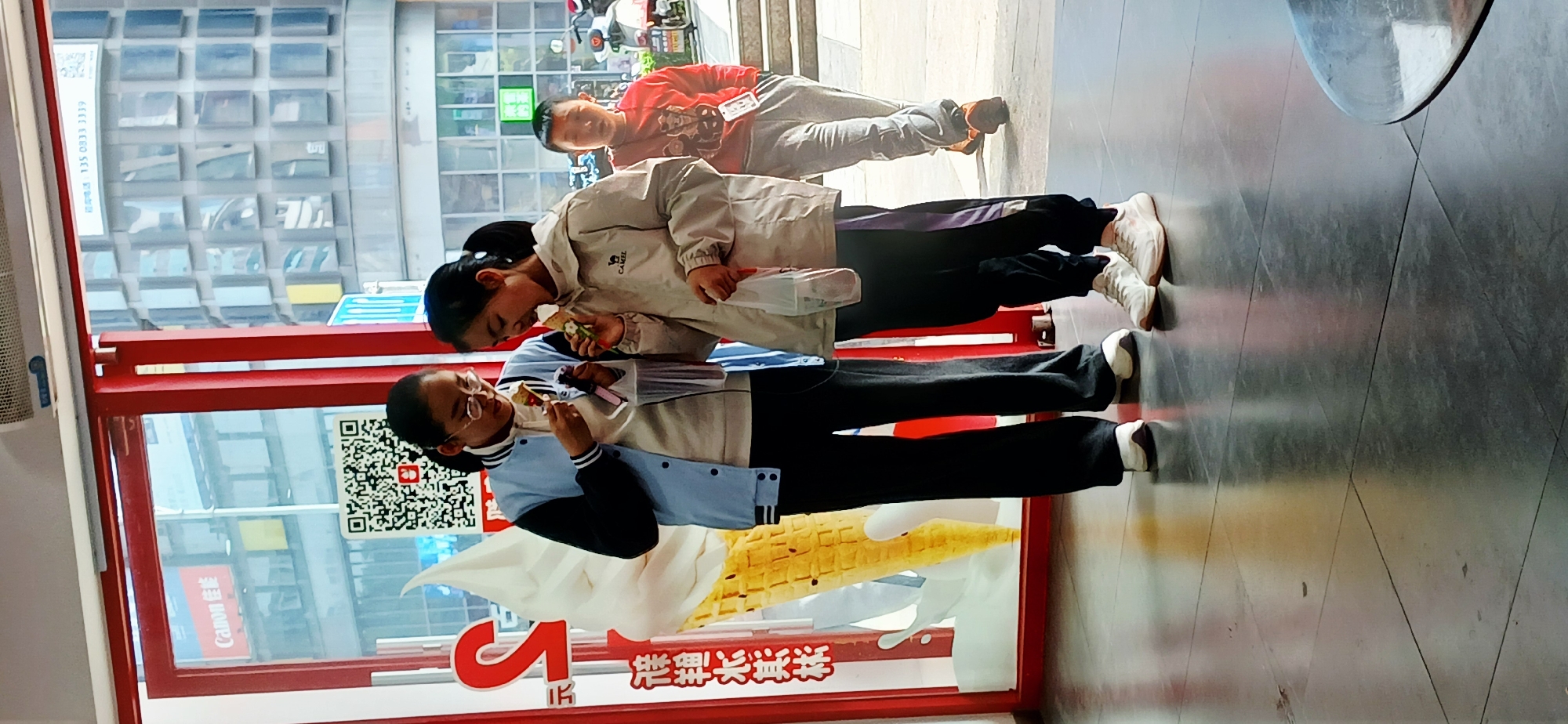 The image depicts three individuals in what appears to be an indoor commercial space. The person closest to the camera is wearing glasses and a blue and white jacket, holding what appears to be an ice cream cone. A second individual in a tan jacket is leaning towards the first, also eating something. A third individual in a red shirt and grey pants is standing further back.
The space has a large advertisement, likely for ice cream, with a QR code visible on the side. There is a window showing a building exterior with advertisements visible. The ground has a reflective surface with geometric patterns.
Based on the Chinese characters on the advertisement and the presence of Chinese individuals, the location is likely in China. There are Chinese Characters indicating Jiulongpo District. Therefore, based on the Chinese characters on the sign and the context of the people, place, and environment, the city is Jiulongpo District, China.