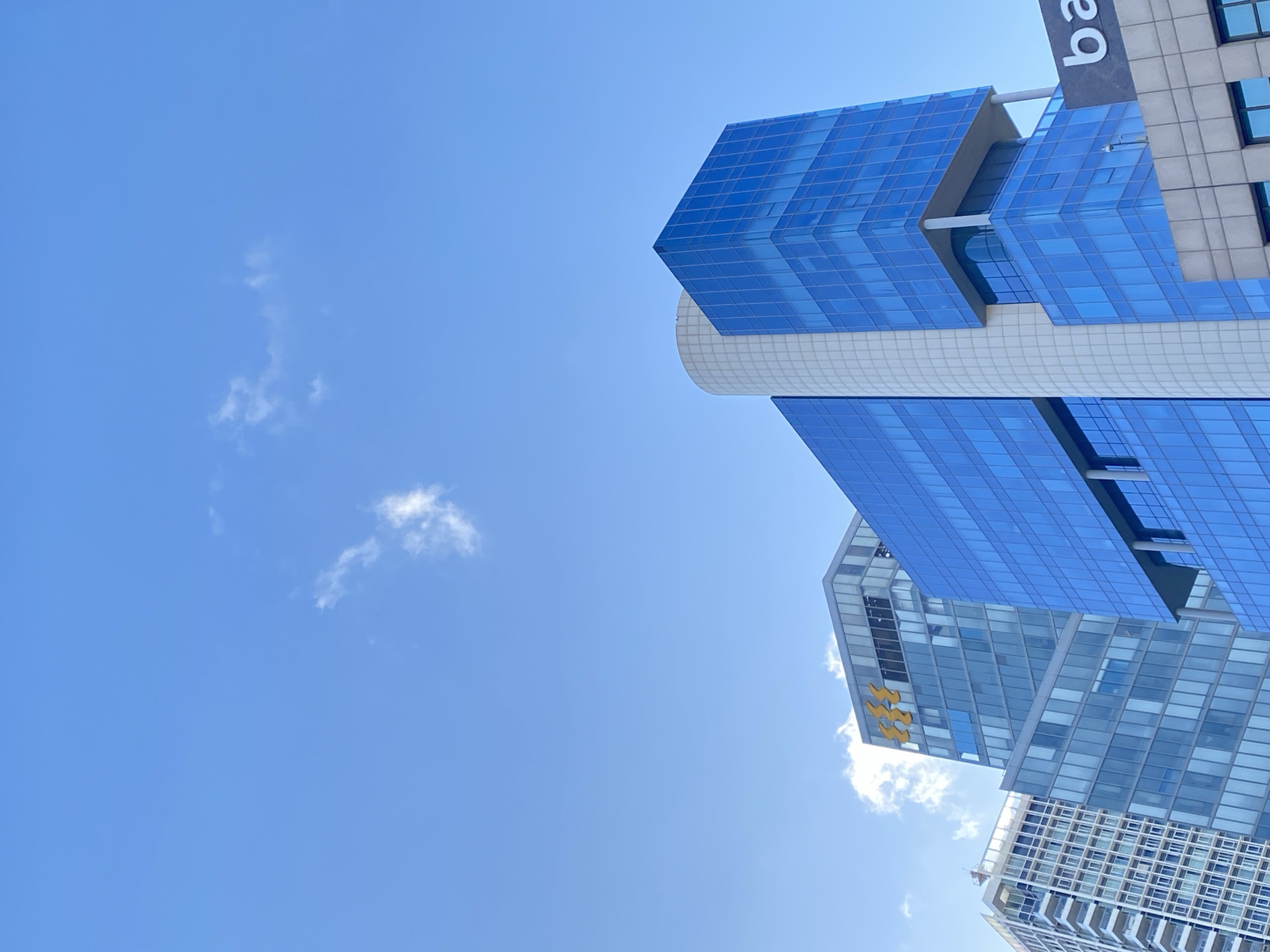 The image depicts a low-angle view of several buildings against a clear, blue sky with sparse clouds. The buildings are located in Tel-Aviv, Israel. The architectural styles appear modern, featuring glass and steel construction. At least one building has a distinguishing architectural feature of a cylindrical column connecting multiple boxy sections. Some buildings are labeled with signs or insignias. No people are visible in the frame.