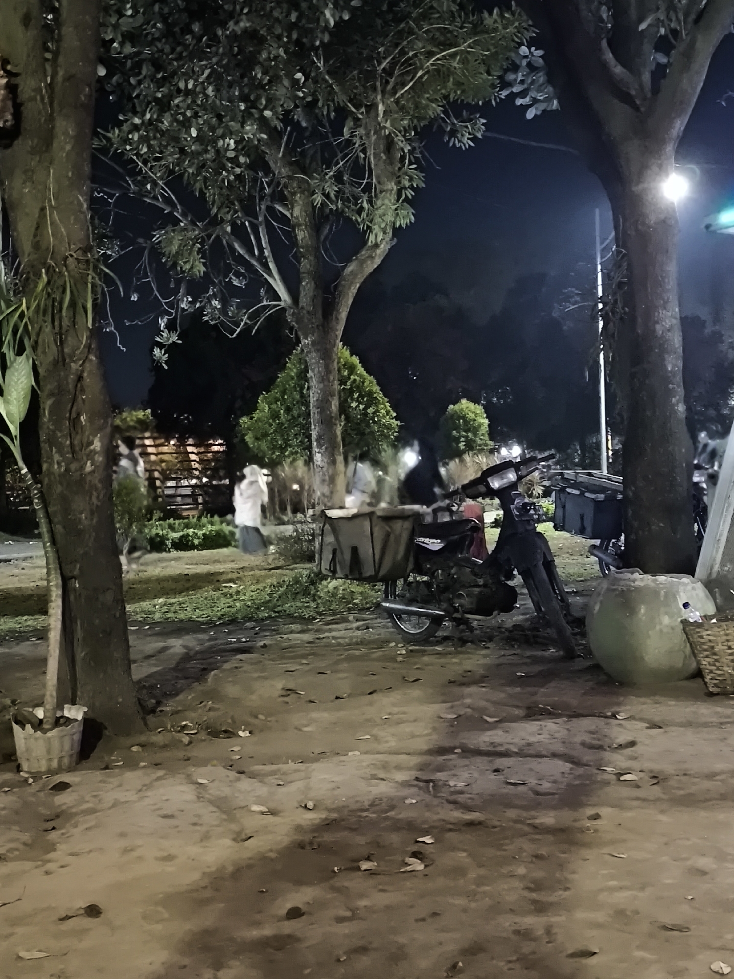 The image depicts an outdoor scene at night, likely in Lumajang, Indonesia, possibly a park or public space, as suggested by the presence of trees and pathways. In the foreground, a motorcycle is parked. It is a dark-colored, older model with what appears to be a basket or cargo carrier attached to the rear. Adjacent to the motorcycle is a large, light-colored planter, and further to the right, a woven basket. The ground is a mixture of dirt and scattered leaves.
In the mid-ground, several figures are visible, appearing somewhat blurred due to the low light conditions and possibly motion. One individual on the left is partially obscured by a tree. Two individuals are more clearly discernible in the center, one wearing a light-colored head covering and garment, and another to their right, also in light clothing. Behind these figures, there are structures that resemble a wooden fence or railing and some greenery.
The background consists of more trees and a dark, night sky. A streetlamp or light fixture is visible to the right, casting illumination on the scene. The overall atmosphere is dim, with artificial lighting contributing to the visibility of the subjects. The presence of the motorcycle and the casual attire of the figures suggest a potentially informal setting.