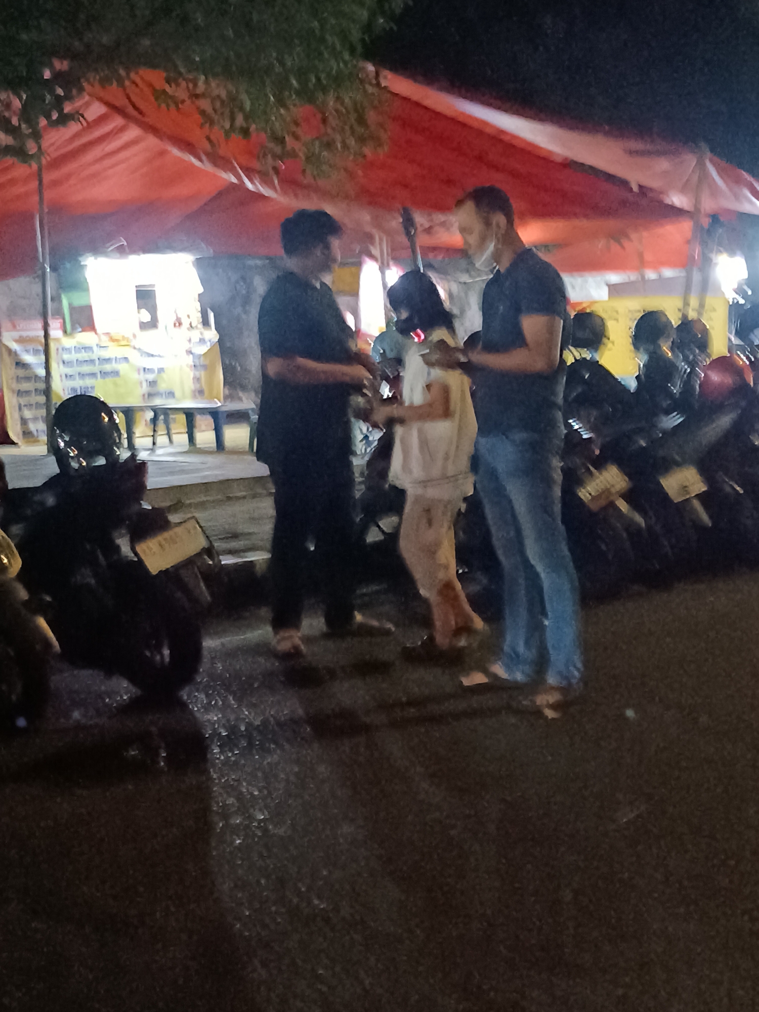 In Yogyakarta, Indonesia, at night, three individuals are standing near a food stall set up under an orange canopy. A man in a dark t-shirt and pants stands on the left, facing away from the camera. To his right, a woman in a light-colored shirt and patterned pants is visible from the side, facing the man on the right. This man, wearing a dark t-shirt and blue jeans, is looking at the woman and holding what appears to be a mobile phone. Behind them, several motorcycles are parked, one with a license plate visible. The ground is asphalt and appears wet or reflective. The scene is illuminated by artificial light, likely from the food stall and surrounding area.