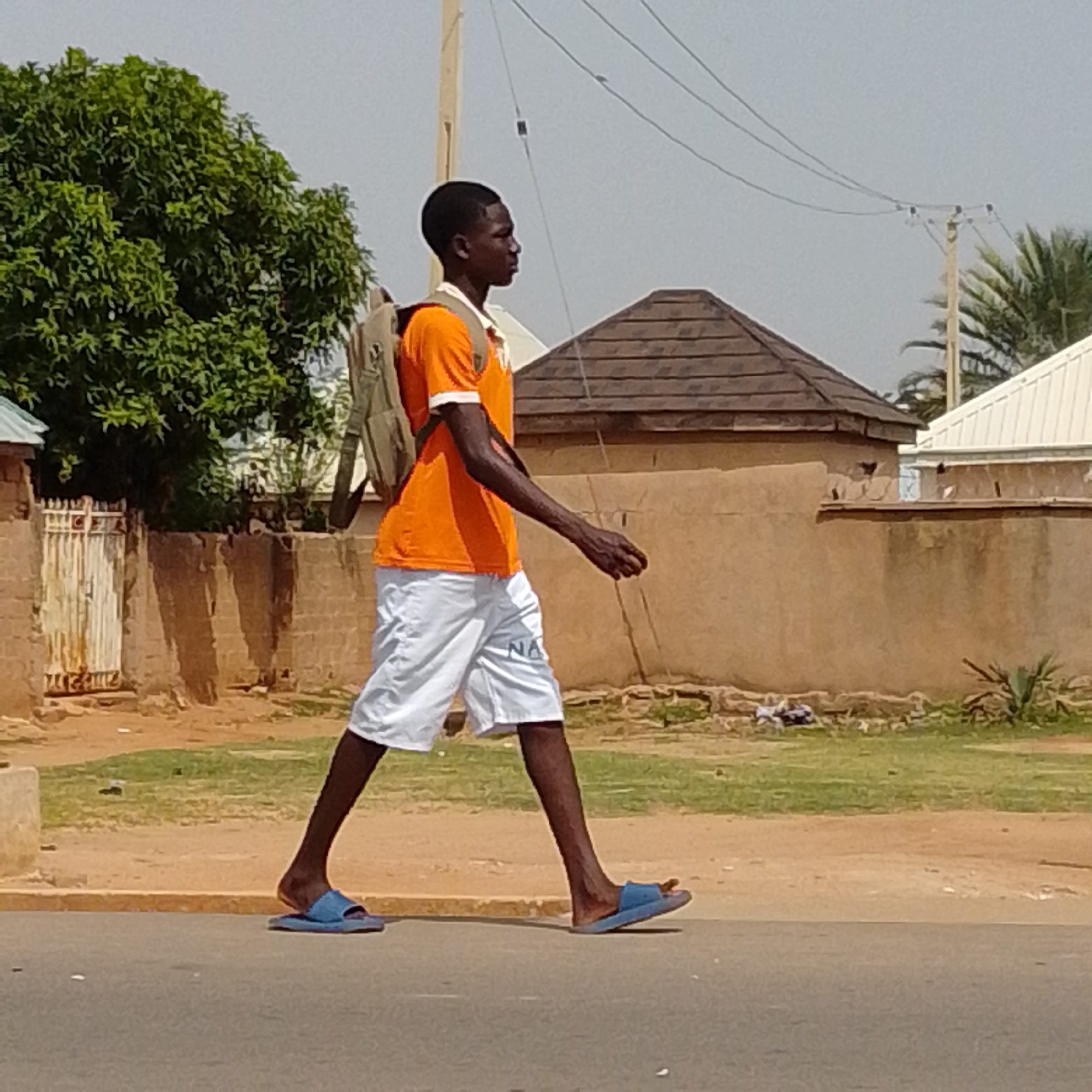 A young Black male is walking from left to right on a paved street in Tempure, Nigeria. He is wearing a bright orange t-shirt with a white collar and trim, and white shorts with the letters "NA" visible on the left leg. He carries a tan backpack over his shoulders and wears blue flip-flops on his feet. The background shows a dirt and grass area with a brick wall topped with a wooden fence. Behind the wall, there are buildings with tiled roofs and a tall wooden utility pole with wires. A large green tree is visible to the left of the frame, and palm fronds can be seen to the right. The sky is a pale blue.