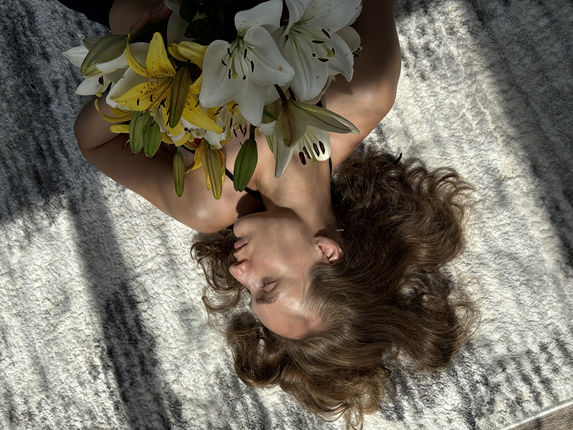 A young woman with long, brown hair lies on her back on a white and grey striped rug, her eyes closed. Sunlight streams across her face and body, creating dappled shadows on the rug. She holds a bouquet of lilies, some white and some yellow with dark speckles, across her chest. Her body is turned to the right, and only her upper body is fully visible. The setting appears to be indoors, possibly in a home in Sharkan, Russia, given the natural lighting and indoor rug.